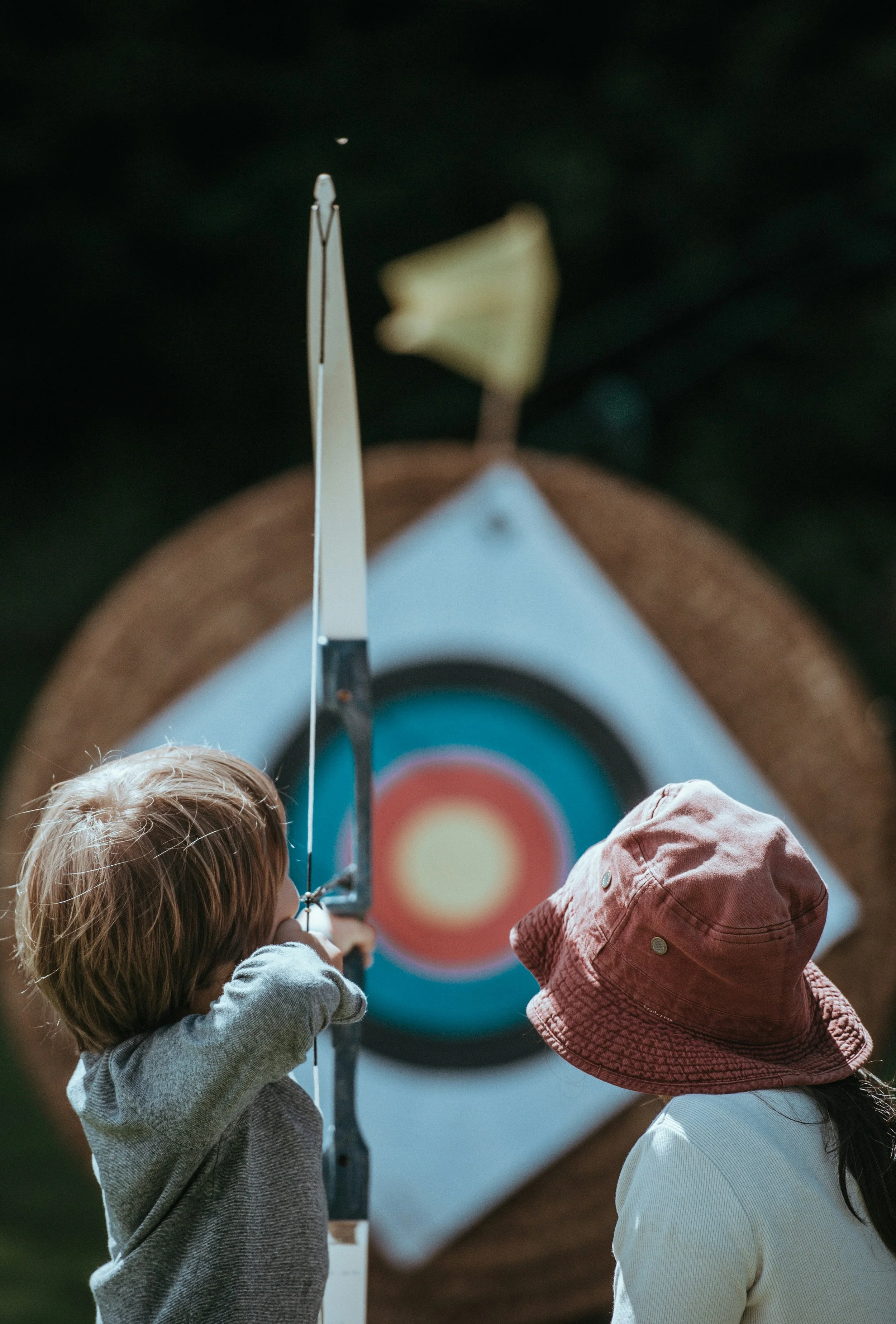 Two children, one with red hair and the other wearing a pink hat, are aiming a bow and arrow at a colorful archery target outdoors.