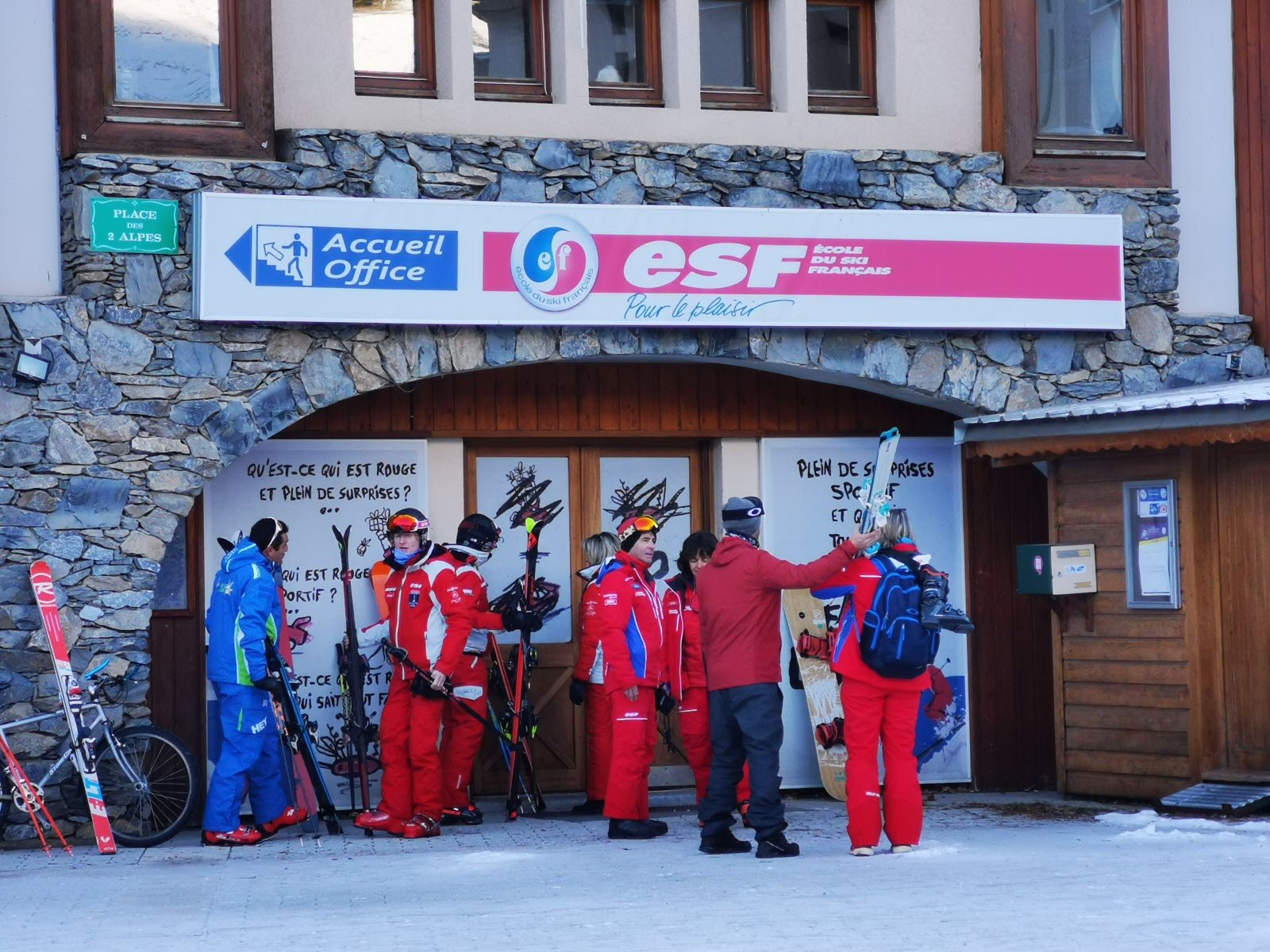 Groupe de personnes en vêtements de ski discutant devant un bâtiment en pierre affichant des panneaux pour l'accueil, le bureau et une école de ski français.