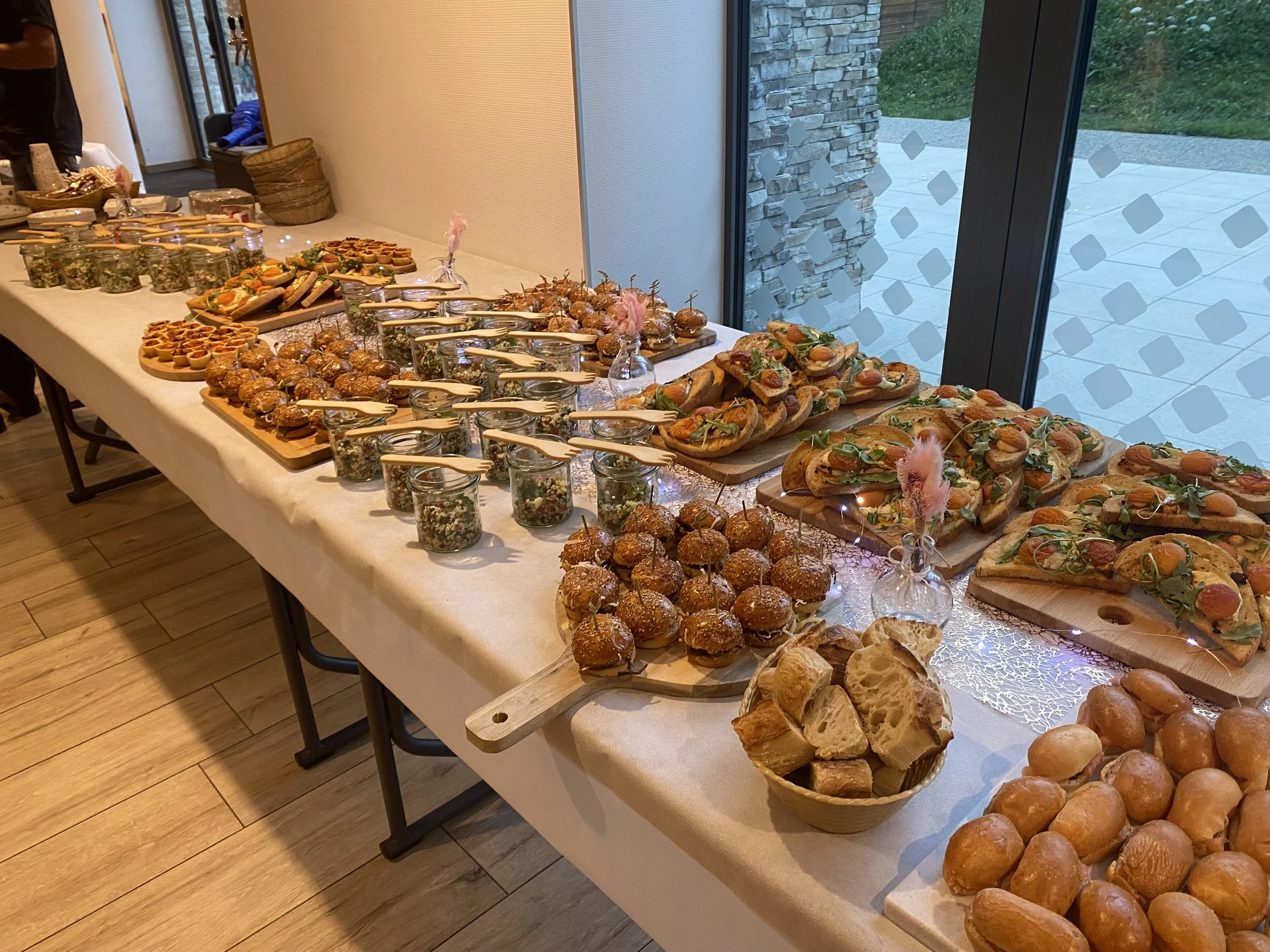 Buffet de plats variés, comprenant des petits sandwiches, des tartines, des mini cakes, des solutions de salade en petits pots et des petits pains, décoré avec des vases contenant des fleurs en plumes. La table est couverte d'une nappe beige clair, dans une pièce lumineuse avec une grande fenêtre donnant sur un espace extérieur.
