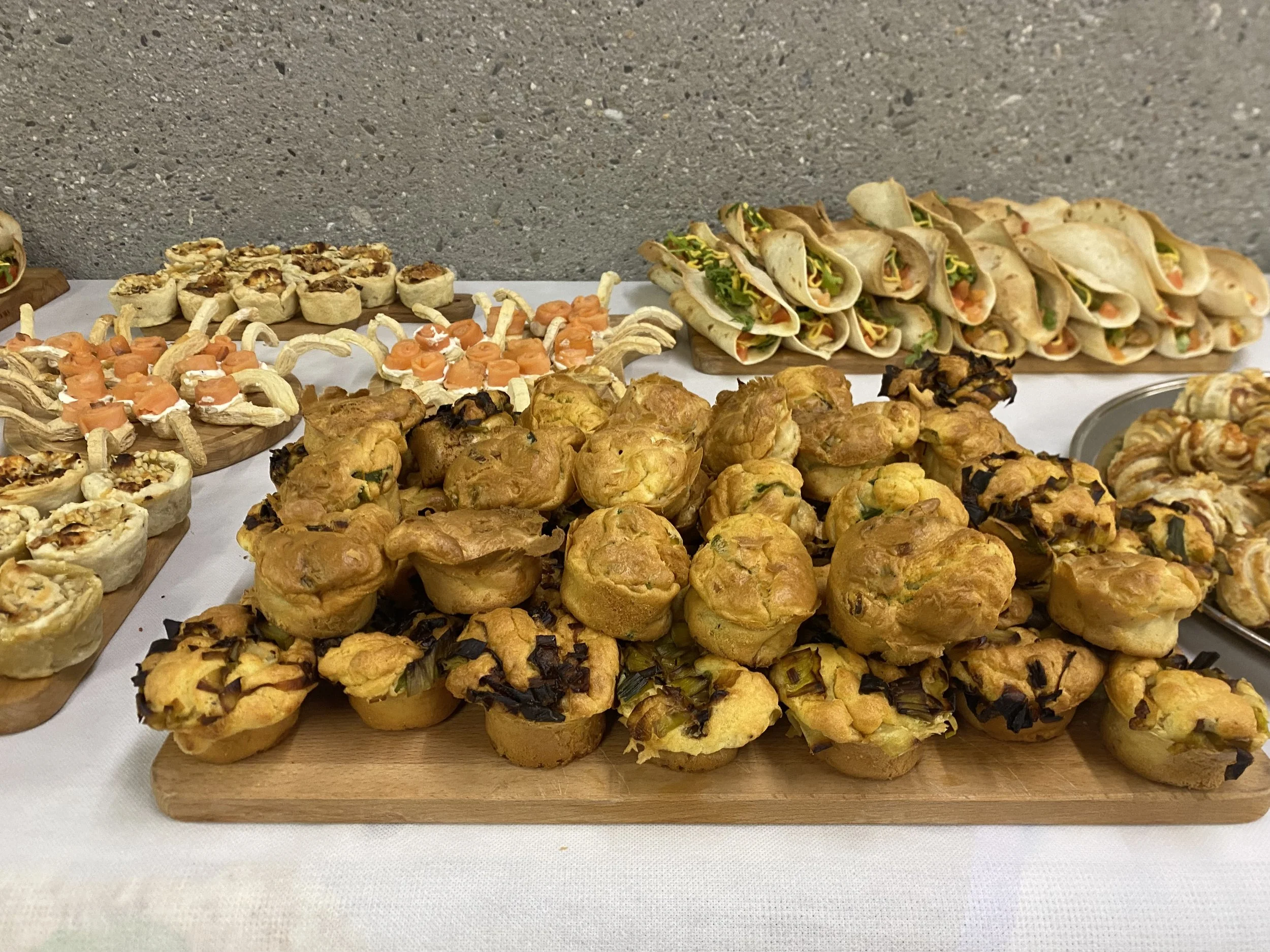 Une table de buffet avec des mini muffins, des bouchées salées, et des wraps. Tout est présenté sur des planches en bois et une assiette en métal visible à droite.