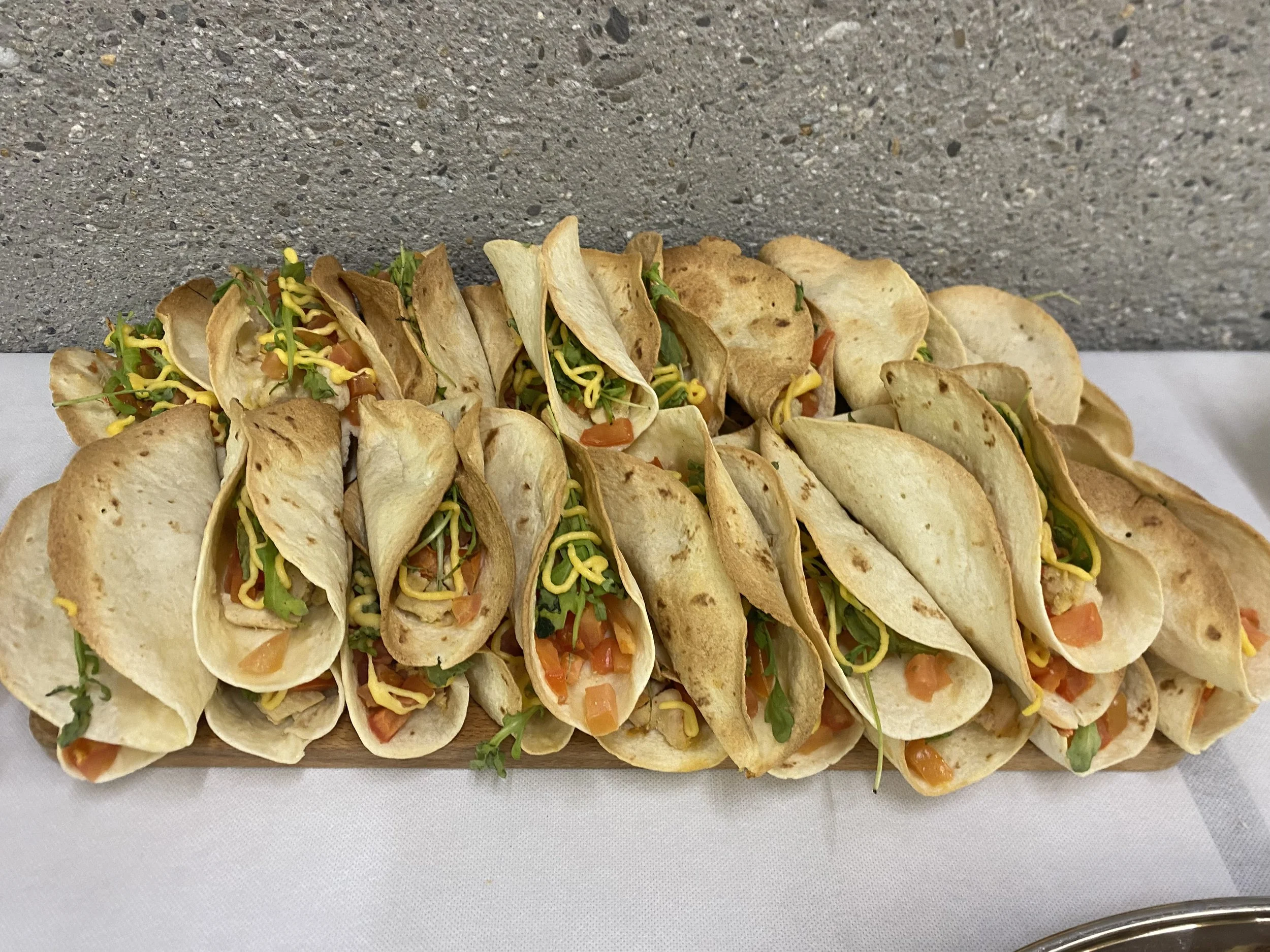 Plateau de tacos avec plusieurs tacos garnis de légumes, fromage et sauce, présenté sur une planche en bois.