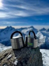 Deux vestes isolantes noires posées sur un rocher avec des montagnes enneigées en arrière-plan.