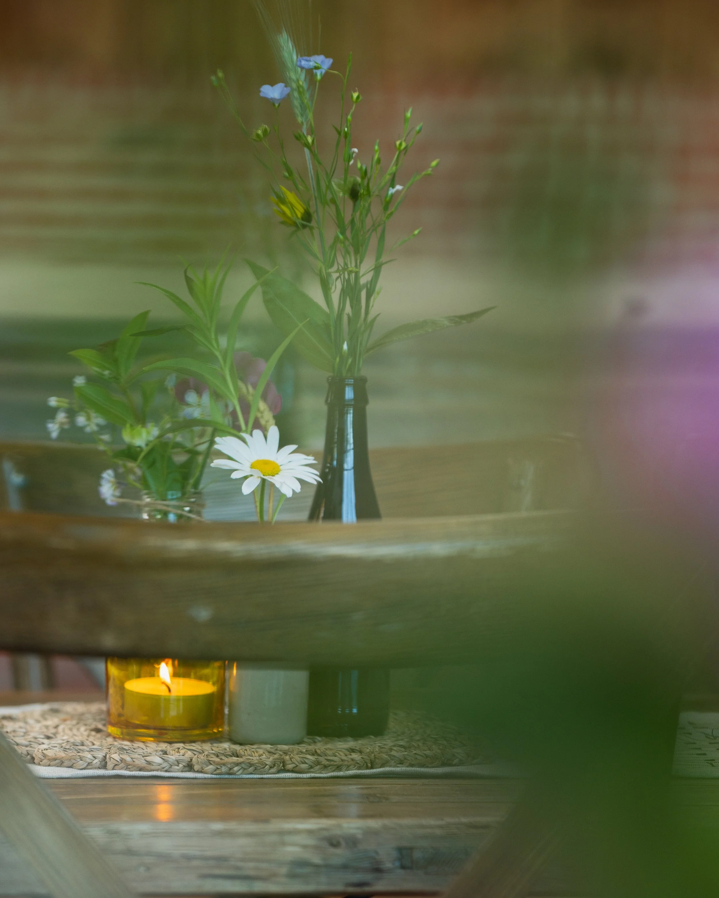 Flowers in a brown bottle and glass, with a lit candle on a wooden table.