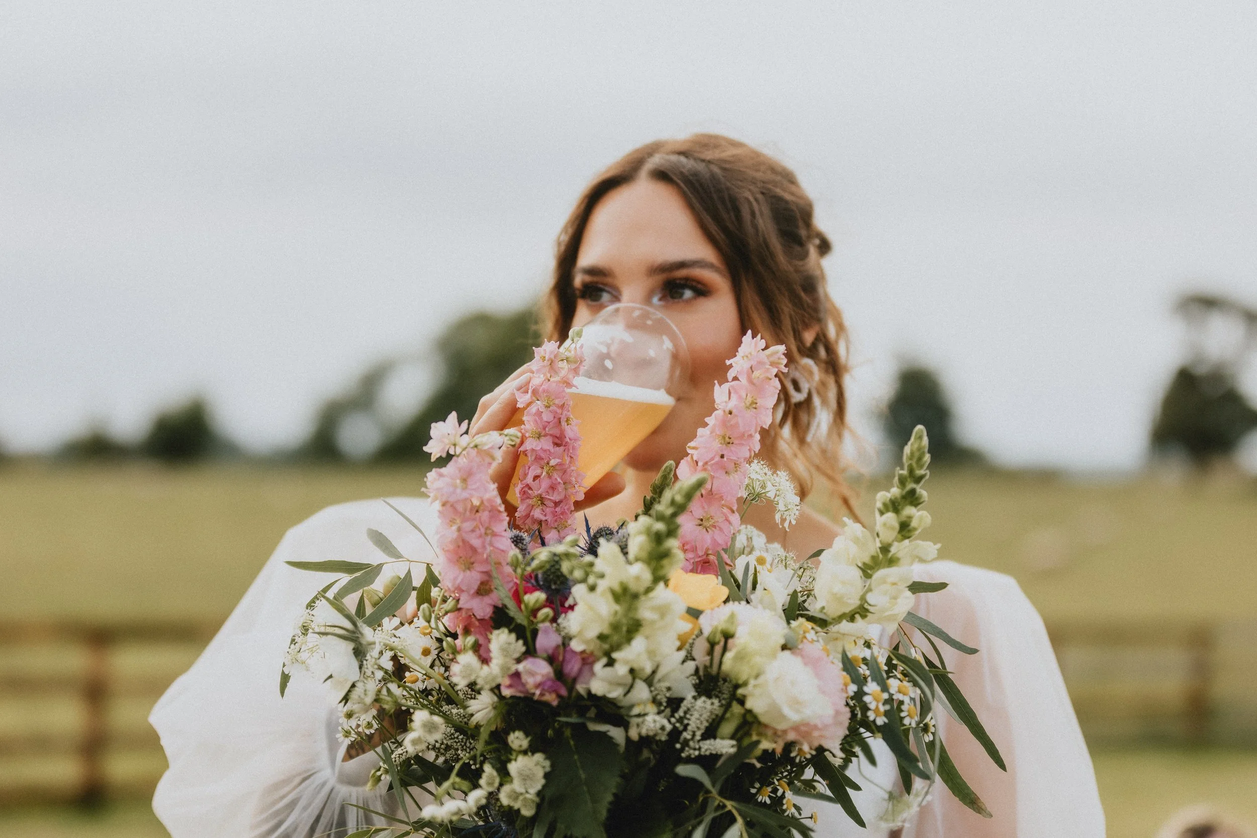 Woman drinking beer behind a bouquet of flowers in an outdoor setting.