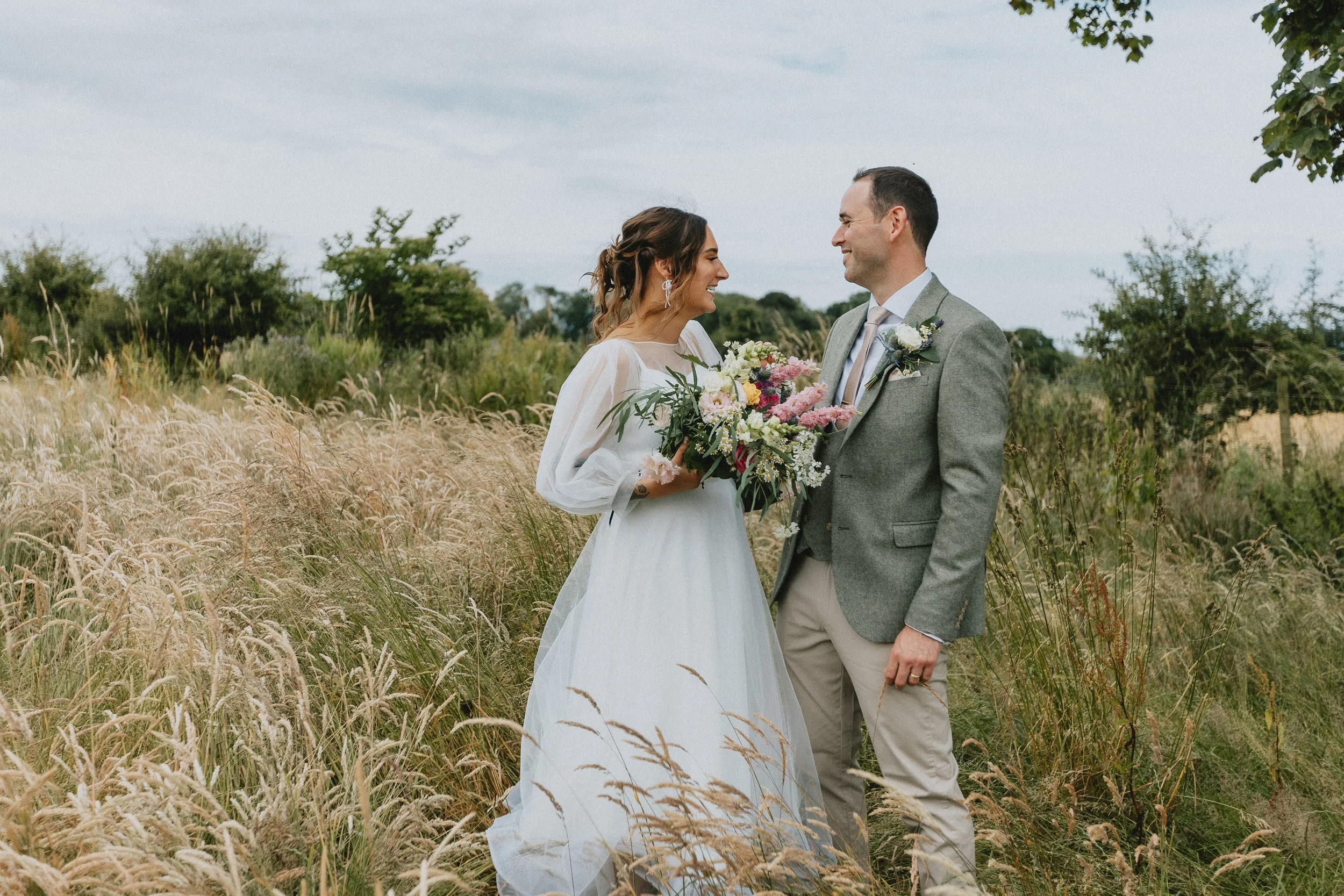 Bride and groom smiling in a field with tall grass and greenery, holding a bouquet of flowers.