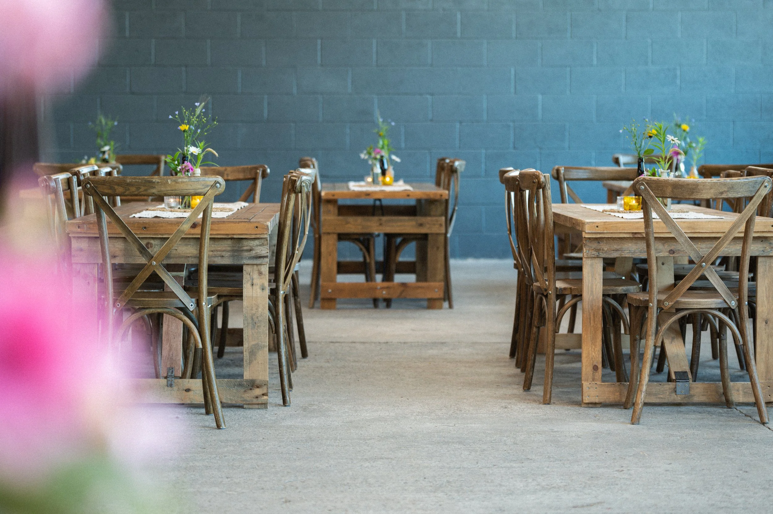 Rustic dining area with wooden tables and chairs, adorned with small floral arrangements and candles, against a blue brick wall.