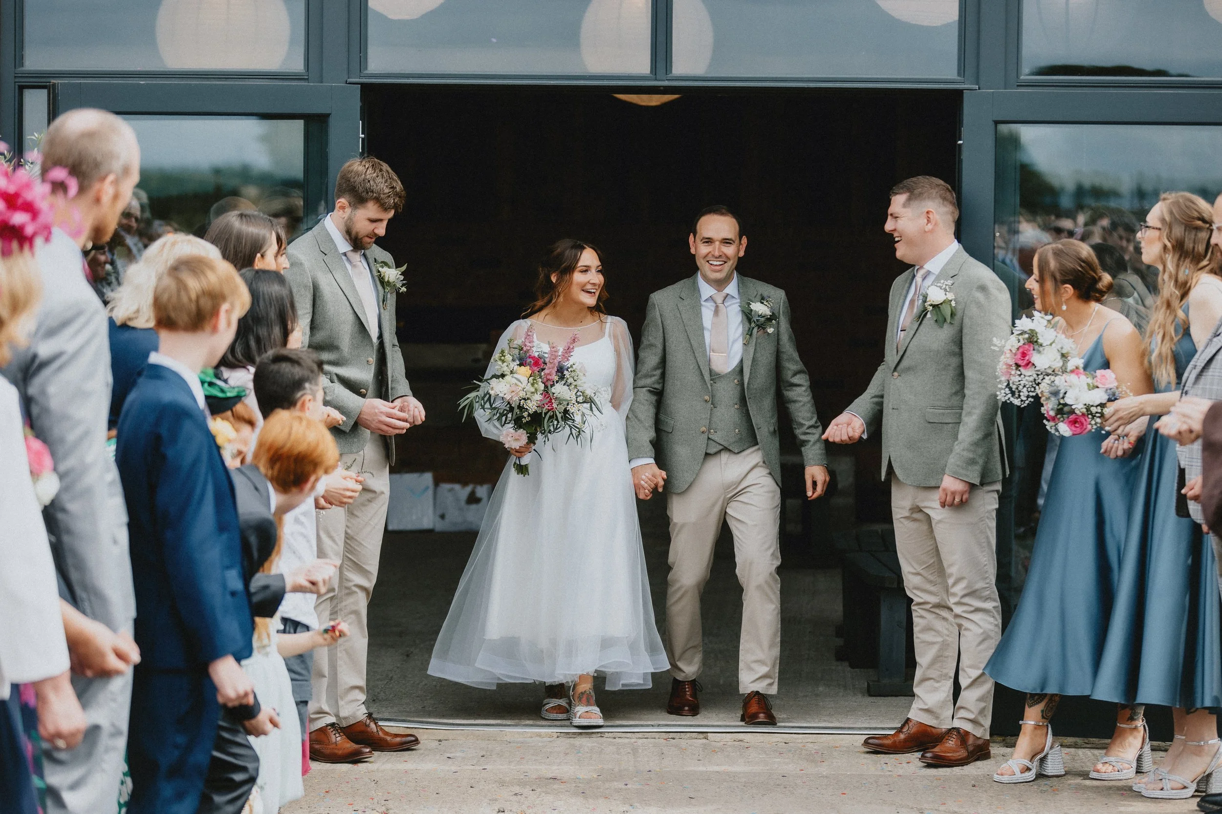 Bride and groom exiting a building, smiling and holding hands, surrounded by guests clapping and holding flowers. The bride holds a bouquet, and groomsmen in green jackets accompany them.