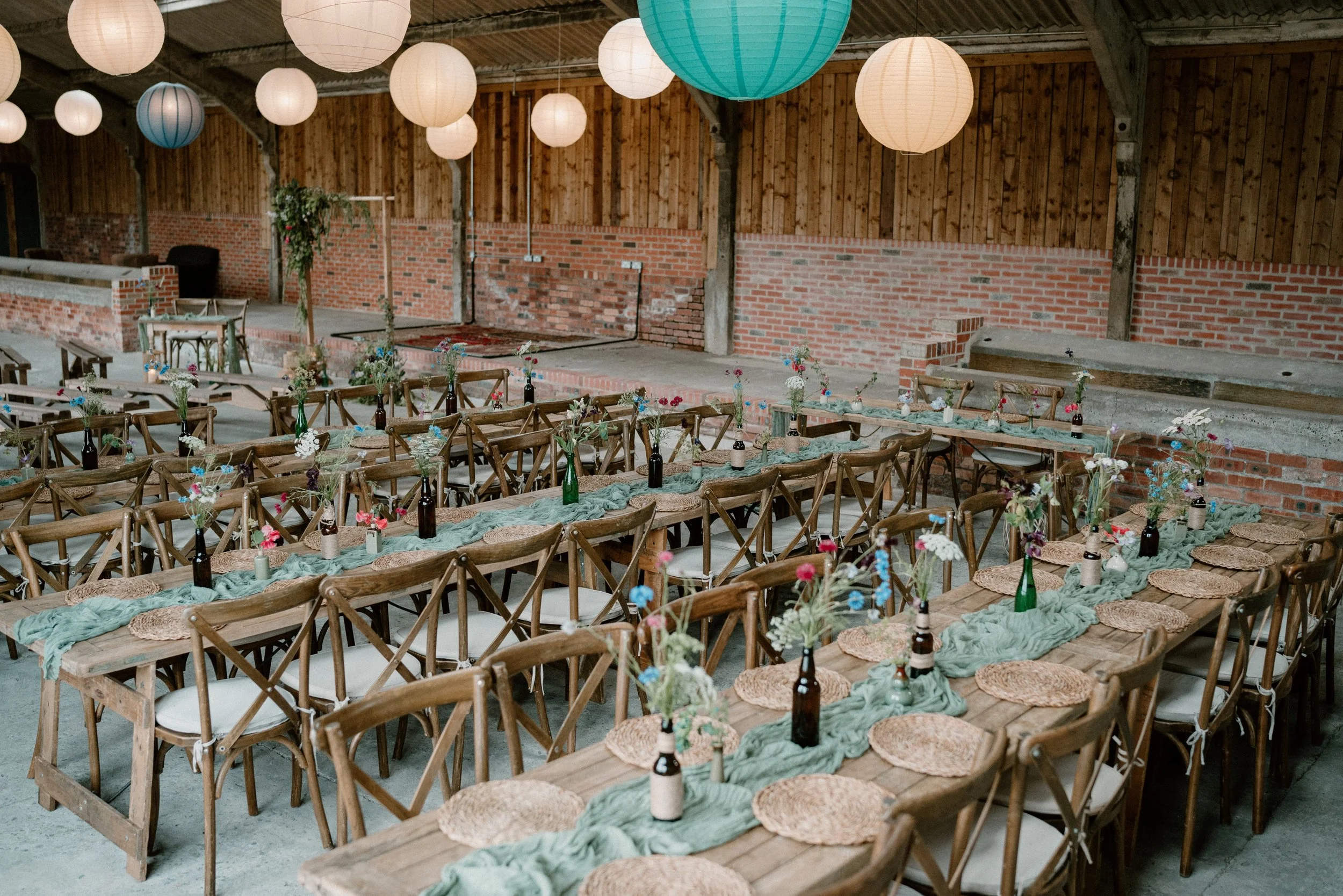 Rustic indoor event setting with wooden tables and chairs, green table runners, wicker placemats, vases with flowers, and hanging paper lanterns.