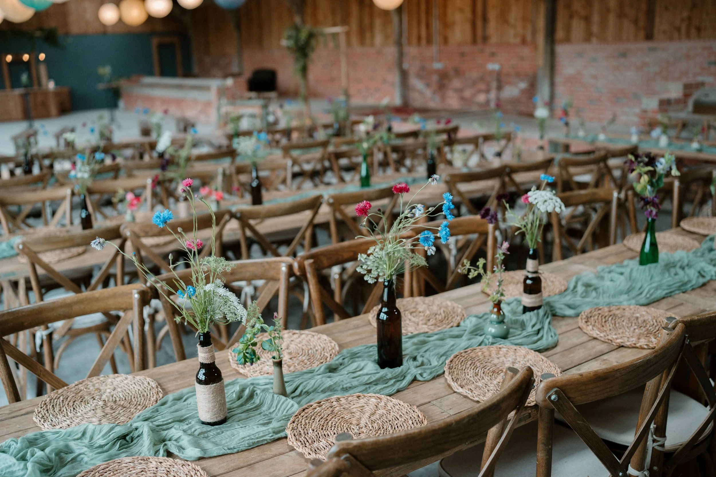Rustic event venue with wooden tables set for dinner, featuring woven placemats, green fabric runners, and bottles with wildflowers as centerpieces, surrounded by wooden chairs.