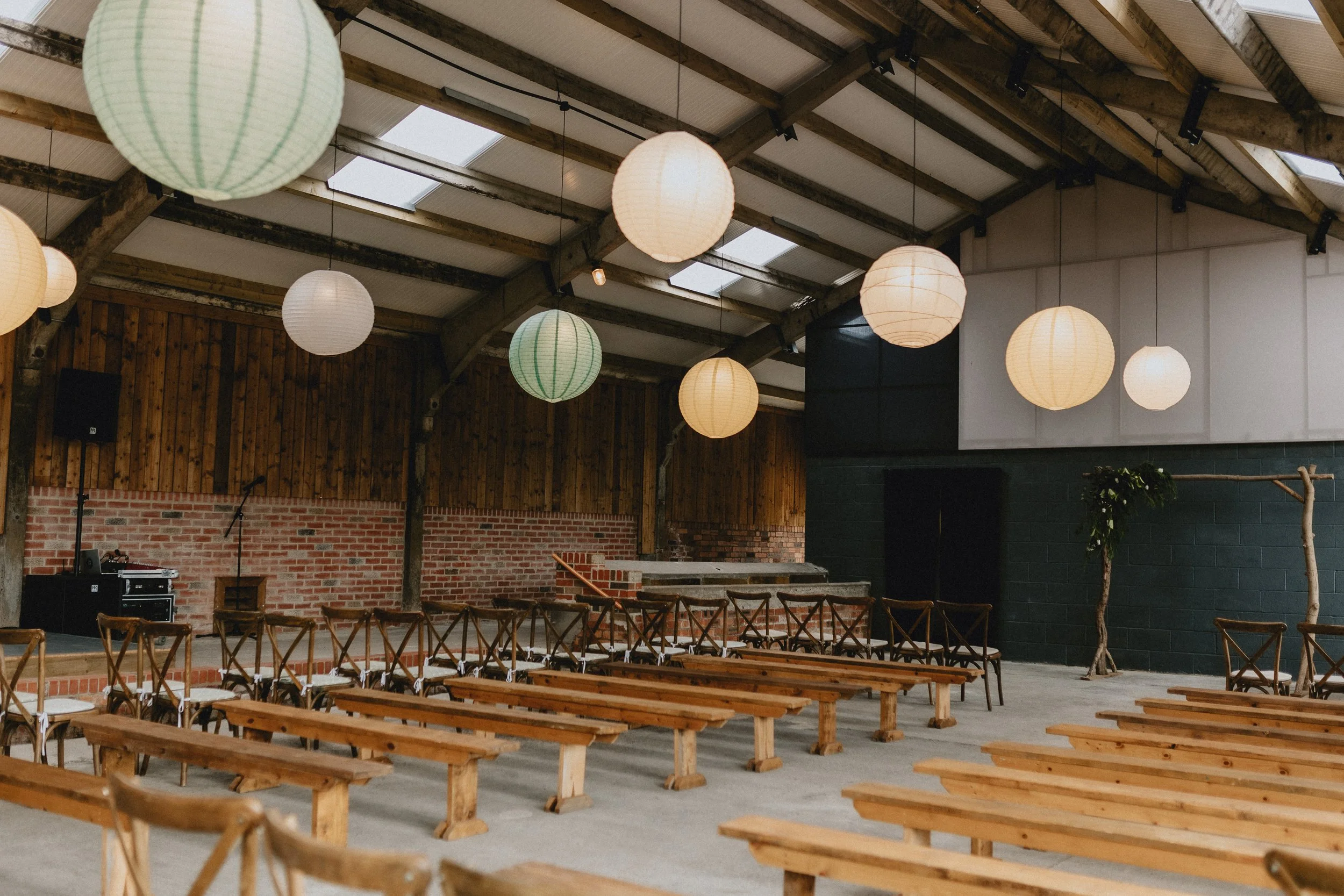 Interior of a rustic event space with wooden benches, paper lanterns hanging from the ceiling, a brick wall, and a stage.
