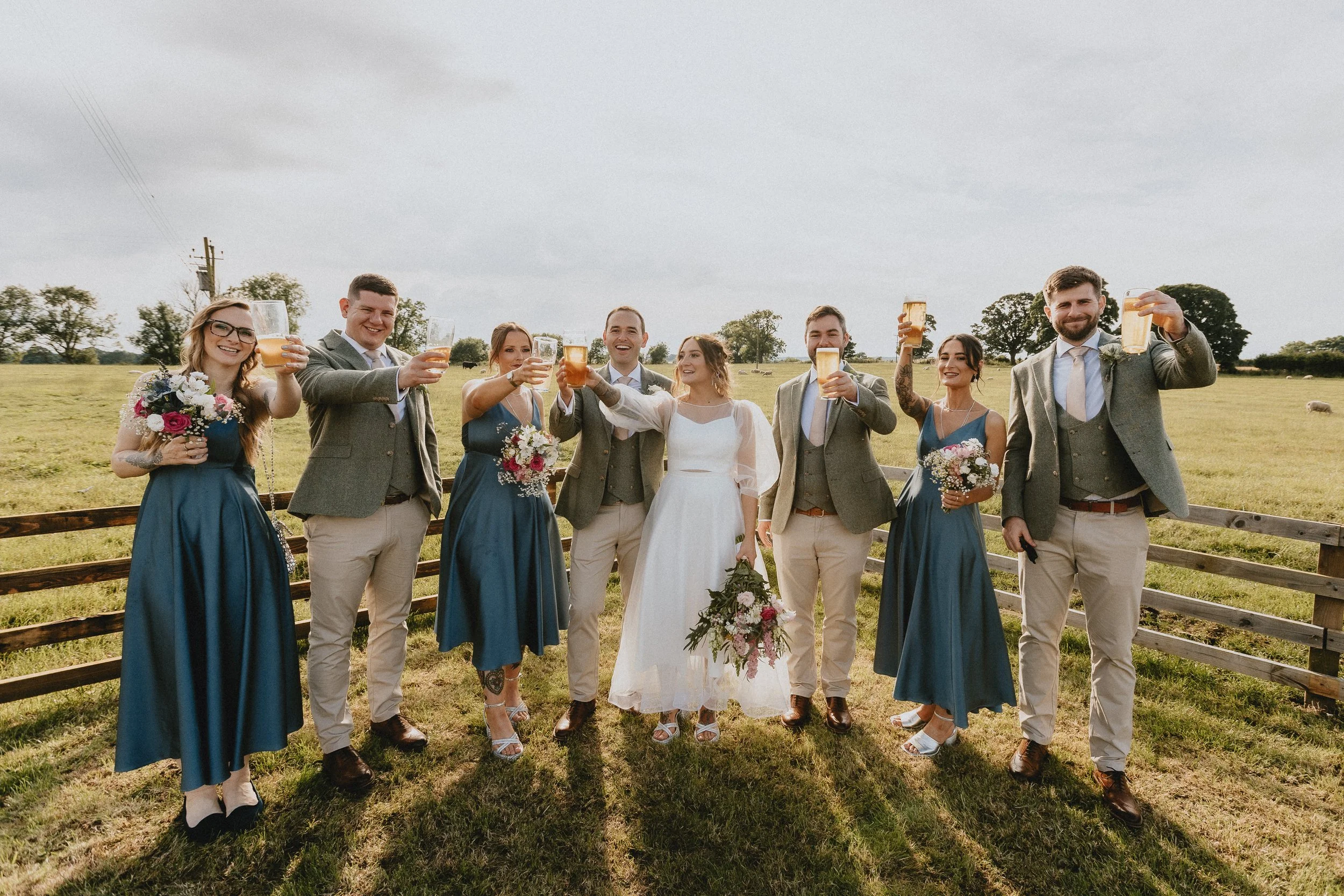 Wedding party raising glasses outdoors, bride in white dress, bridesmaids in blue, groomsmen in suits, countryside setting.