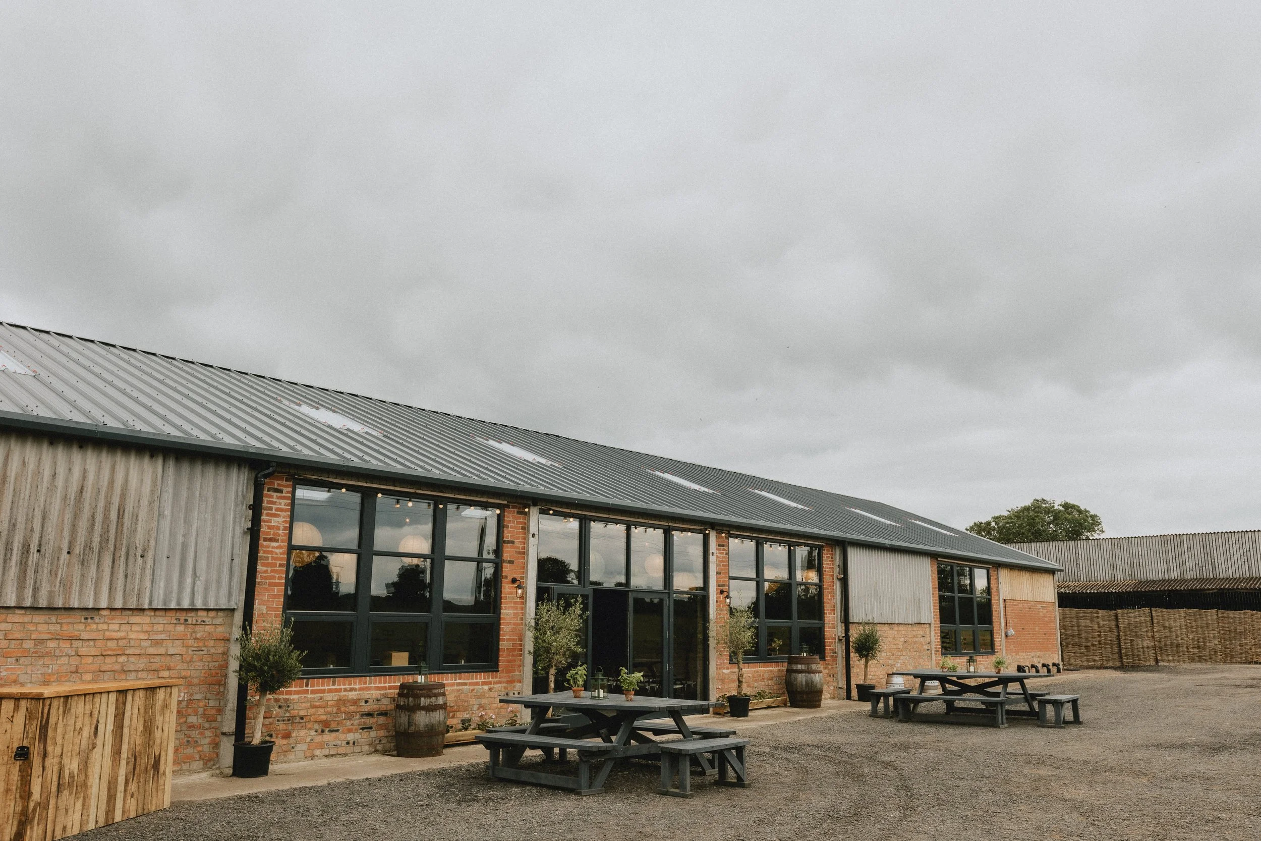 Industrial-style building with brick and tin exterior, featuring large windows and picnic tables outside on a gravel surface. Cloudy sky background.