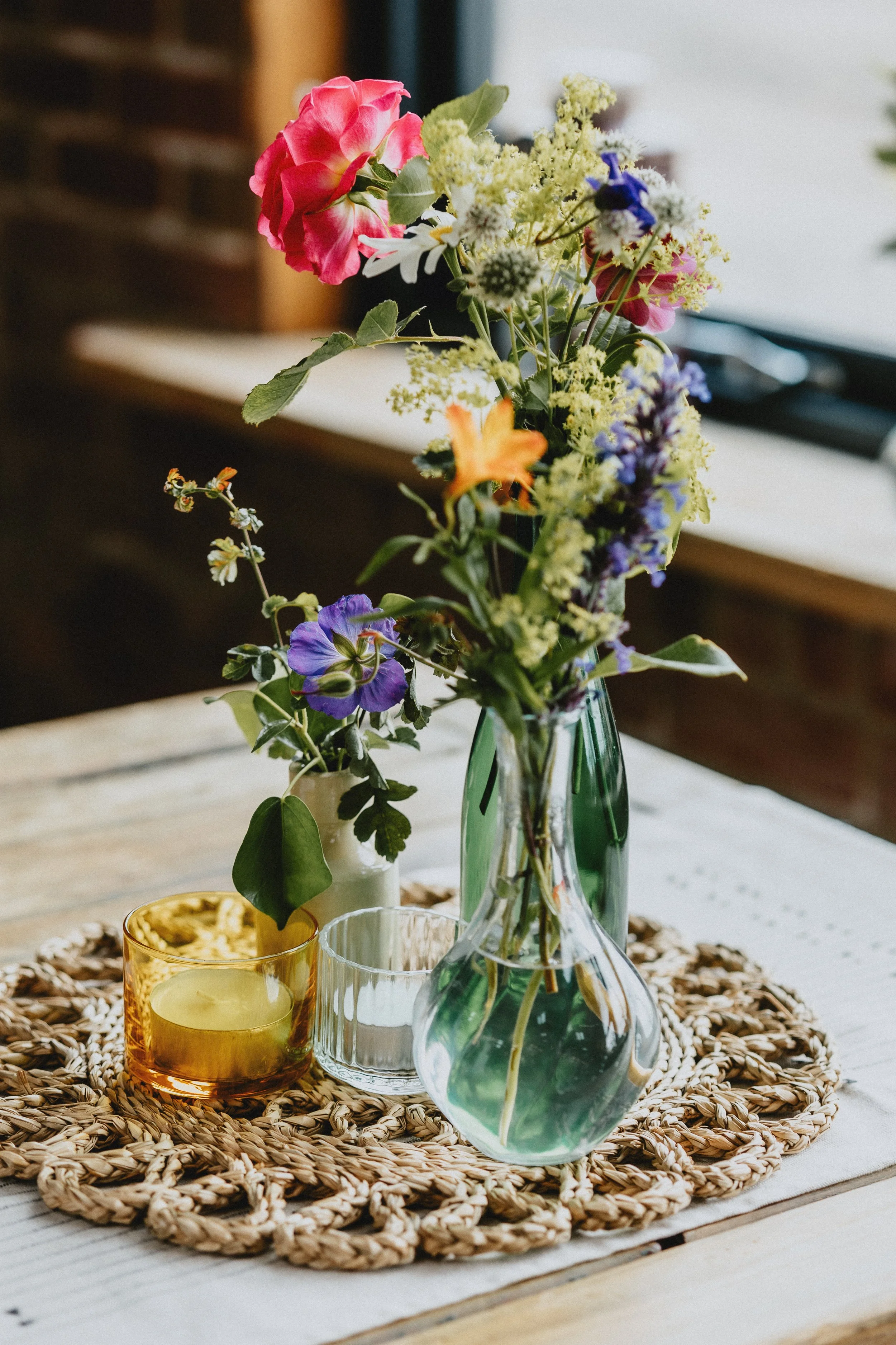 A rustic table setting with a glass vase holding a bouquet of colorful flowers including pink, yellow, and purple blooms, placed on a woven placemat. Two small candles in glass holders are nearby.