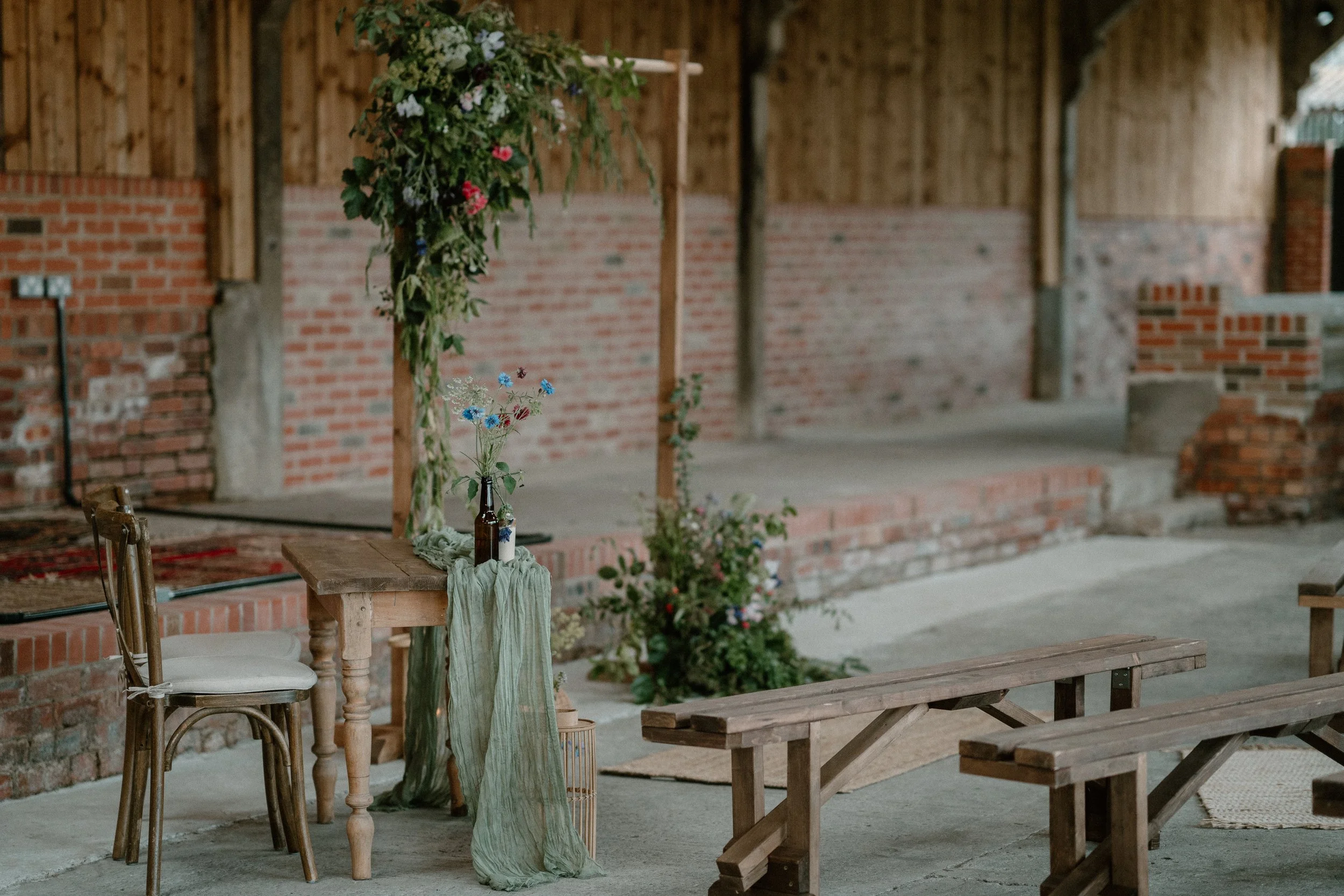 Rustic indoor setting with a wooden table and chair, decorated with a greenery arch and wildflowers, next to wooden benches and a brick wall.
