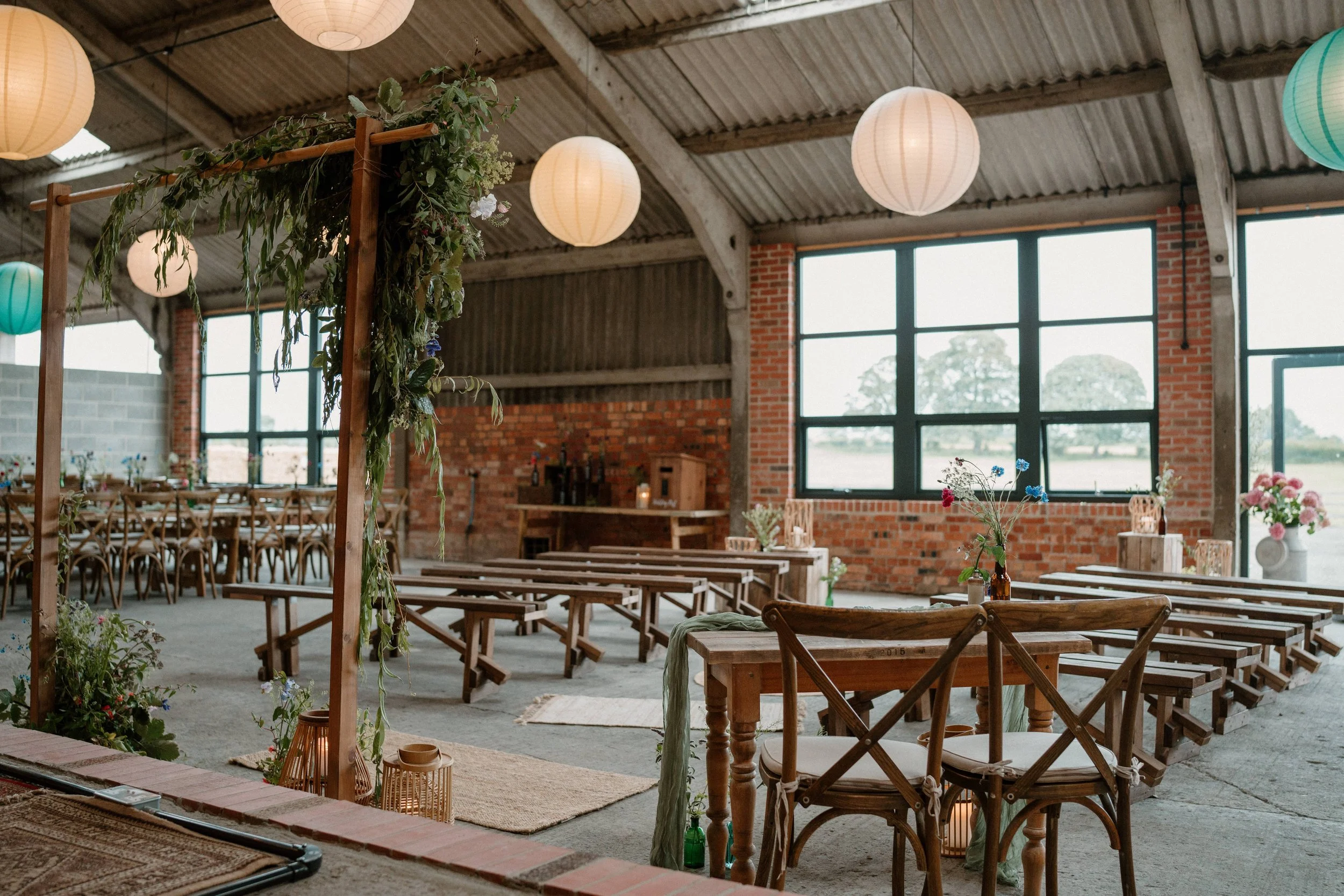 Rustic barn wedding setup with wooden benches and tables, hanging paper lanterns, brick walls, and greenery decorations.