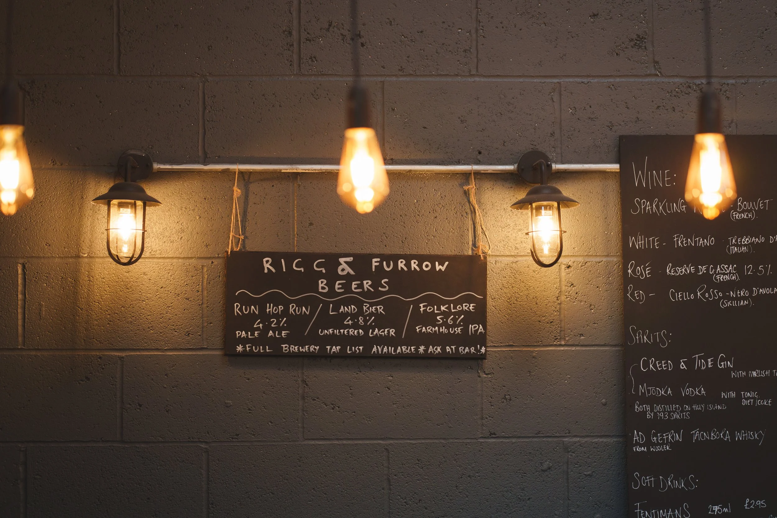 Cafe menu on a wall with industrial-style lights, featuring a chalkboard listing beers such as 'Run Hop Run,' 'Land Bier,' and 'Folklore.' Another board lists wines and spirits.