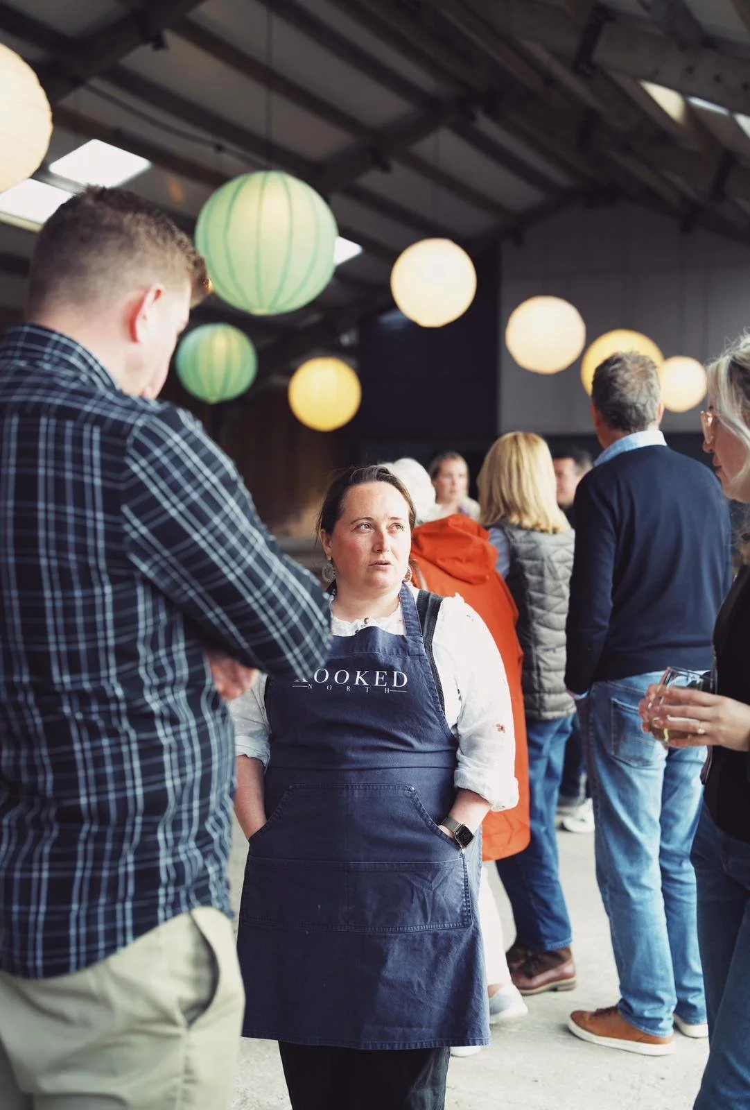 Group of people socializing indoors with paper lanterns overhead, focus on individual wearing an apron.