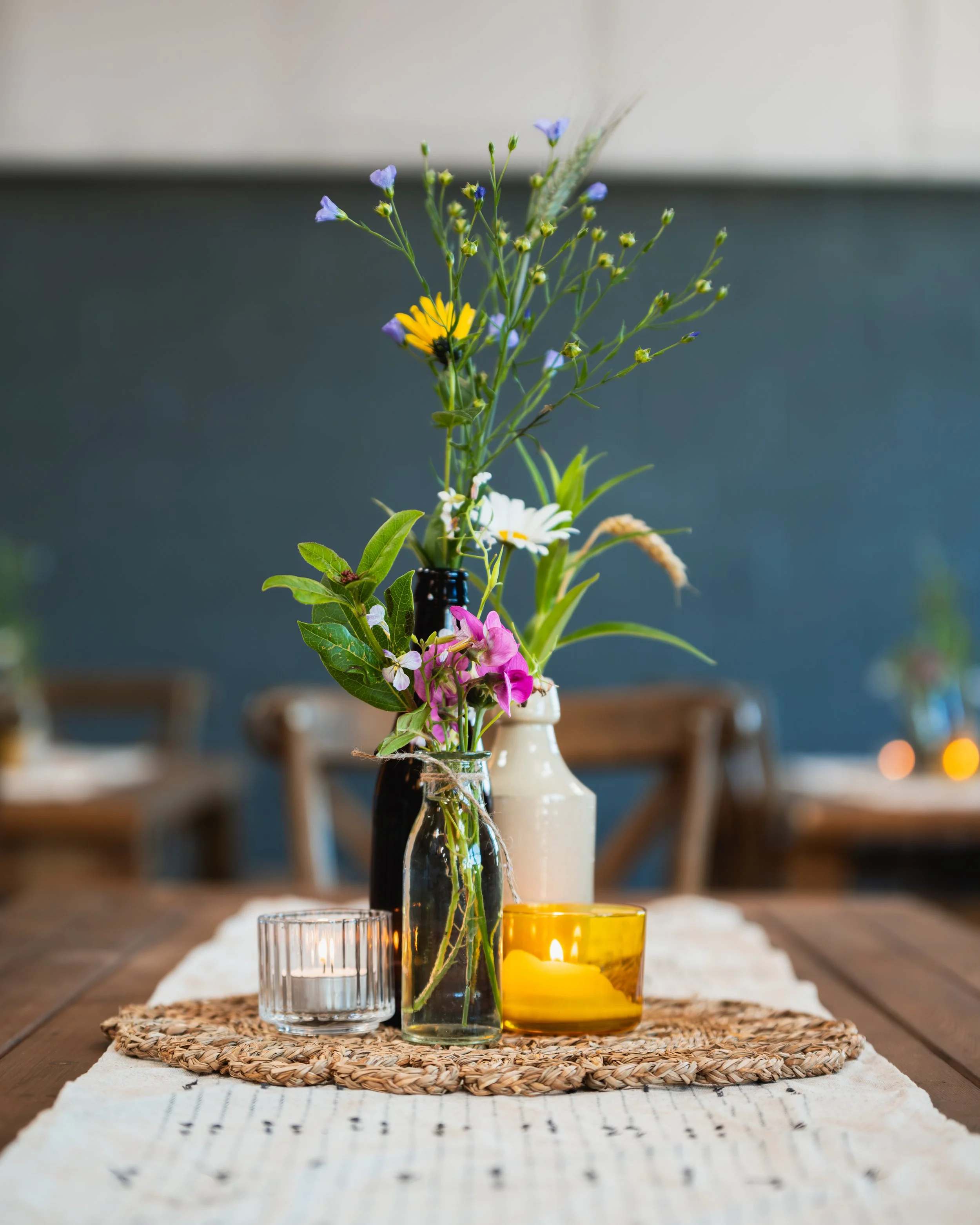 A wooden table with a floral centerpiece including wildflowers in glass vases, two lit candles in holders, and a woven placemat.
