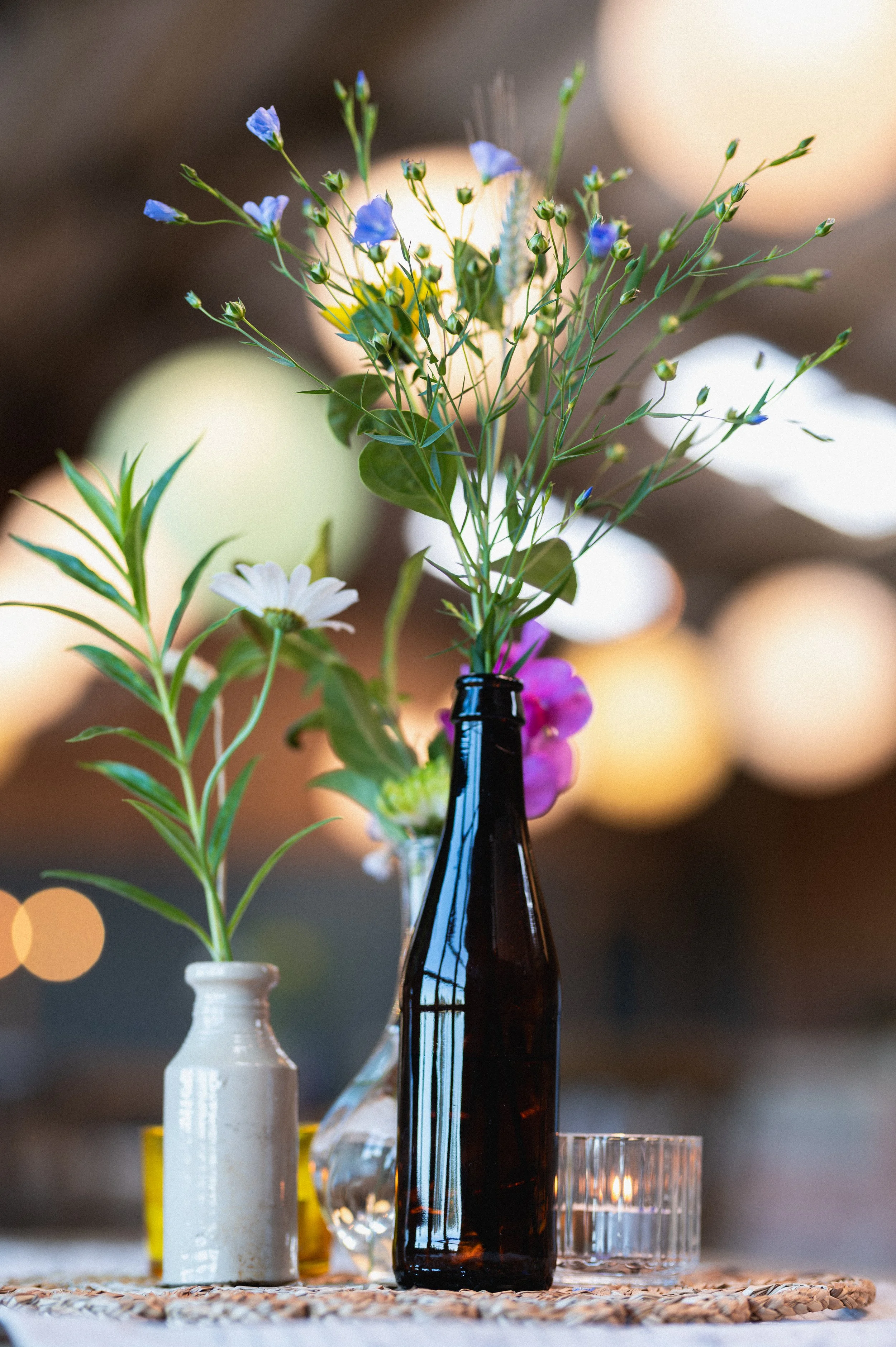 Vases with flowers on a table, including a brown bottle with wildflowers, a smaller vase with a white daisy, and another with blurred blooms in the background, set against a warm, blurred backdrop.