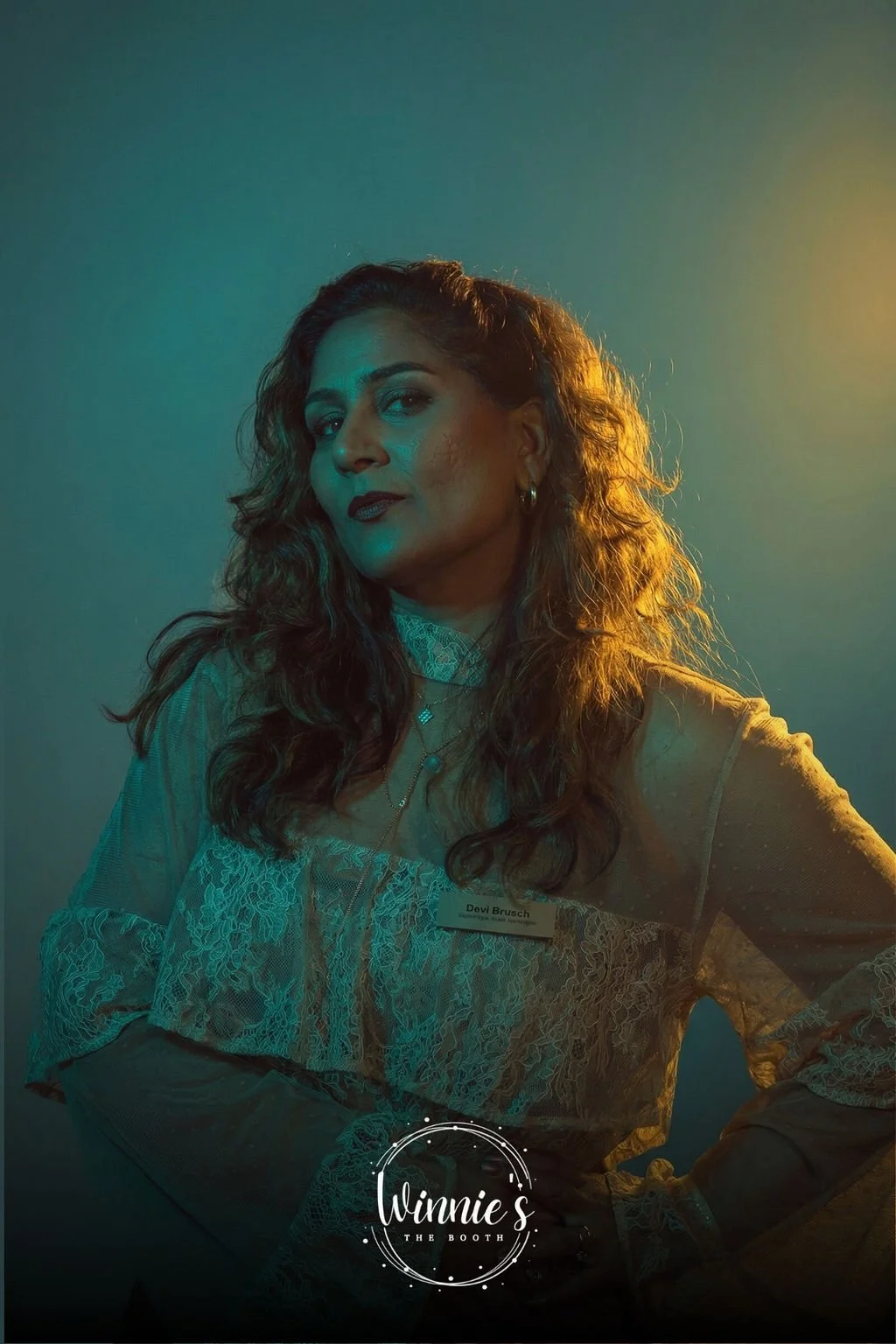 A group of five people, three women and two men, posing together in a cozy, elegant indoor setting with floral decorations and a large abstract painting behind them. All are dressed in formal or semi-formal attire, with two women wearing blue dresses and the man standing in a black suit with a pink tie.