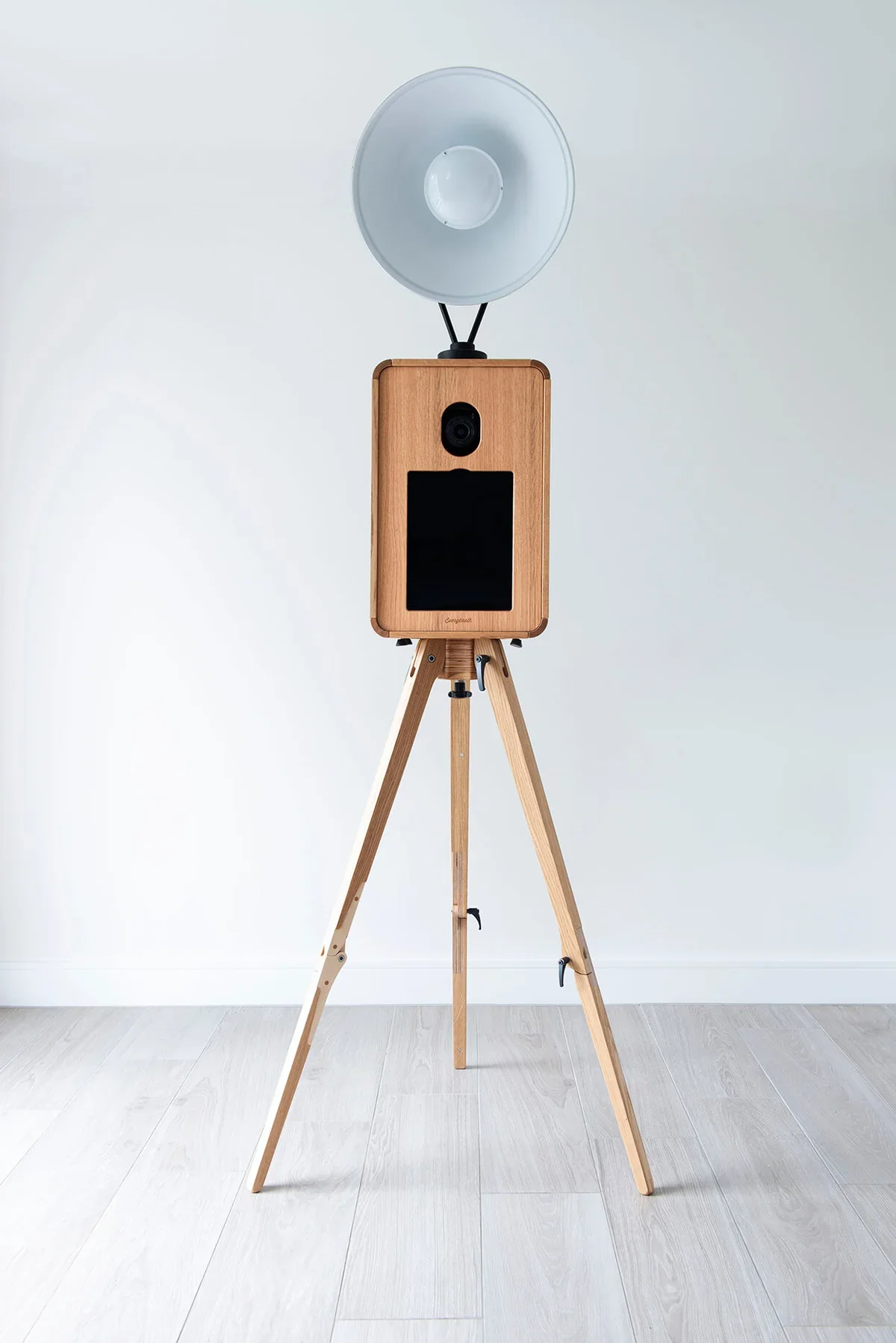 A vintage wooden camera with a large white flash attachment on a tripod against a plain white wall.