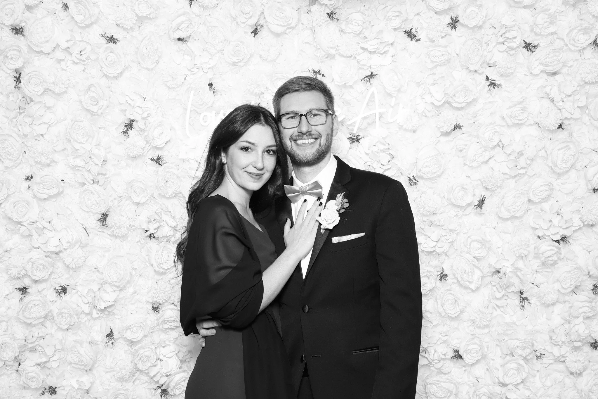 A man in a black suit and tie kisses a smiling woman wearing a black dress and earrings.