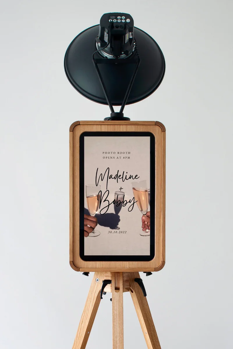 Photo booth with a digital screen displaying a wedding invitation for Madeline and Bobby, dated 30.10.2022, on a wooden tripod stand, with a black photo booth device mounted above.