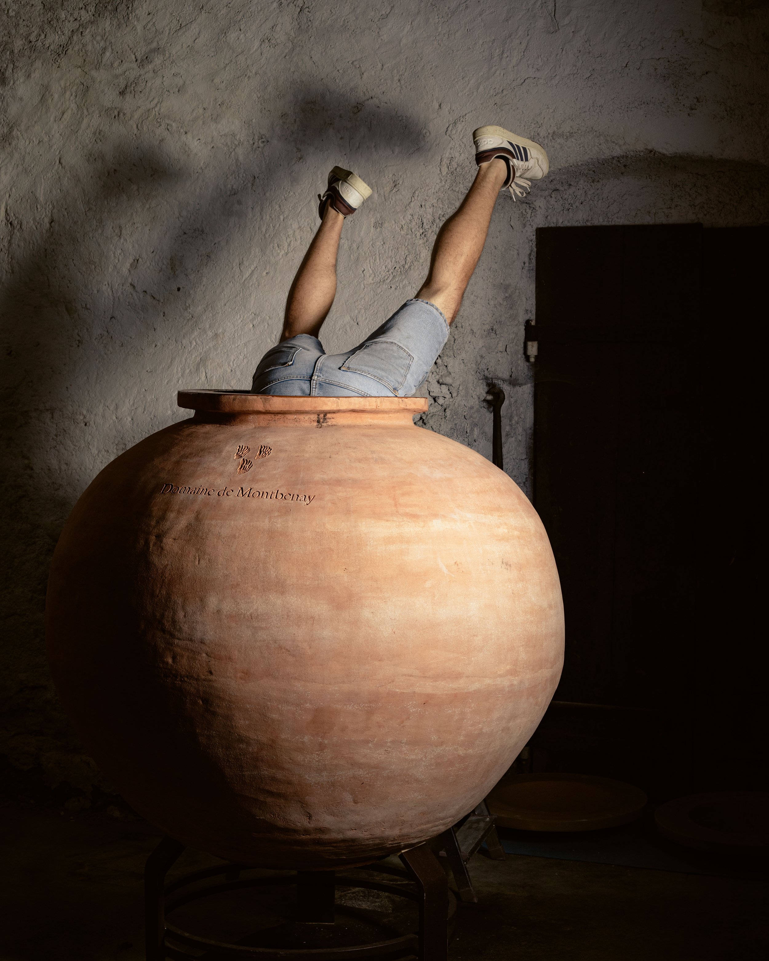 Une personne en shorts et baskets, dont seules les jambes et une partie du tronc sont visibles, est allongée dans une grande jarre en terre cuite, avec ses jambes levées en l'air, dans une pièce sombre.