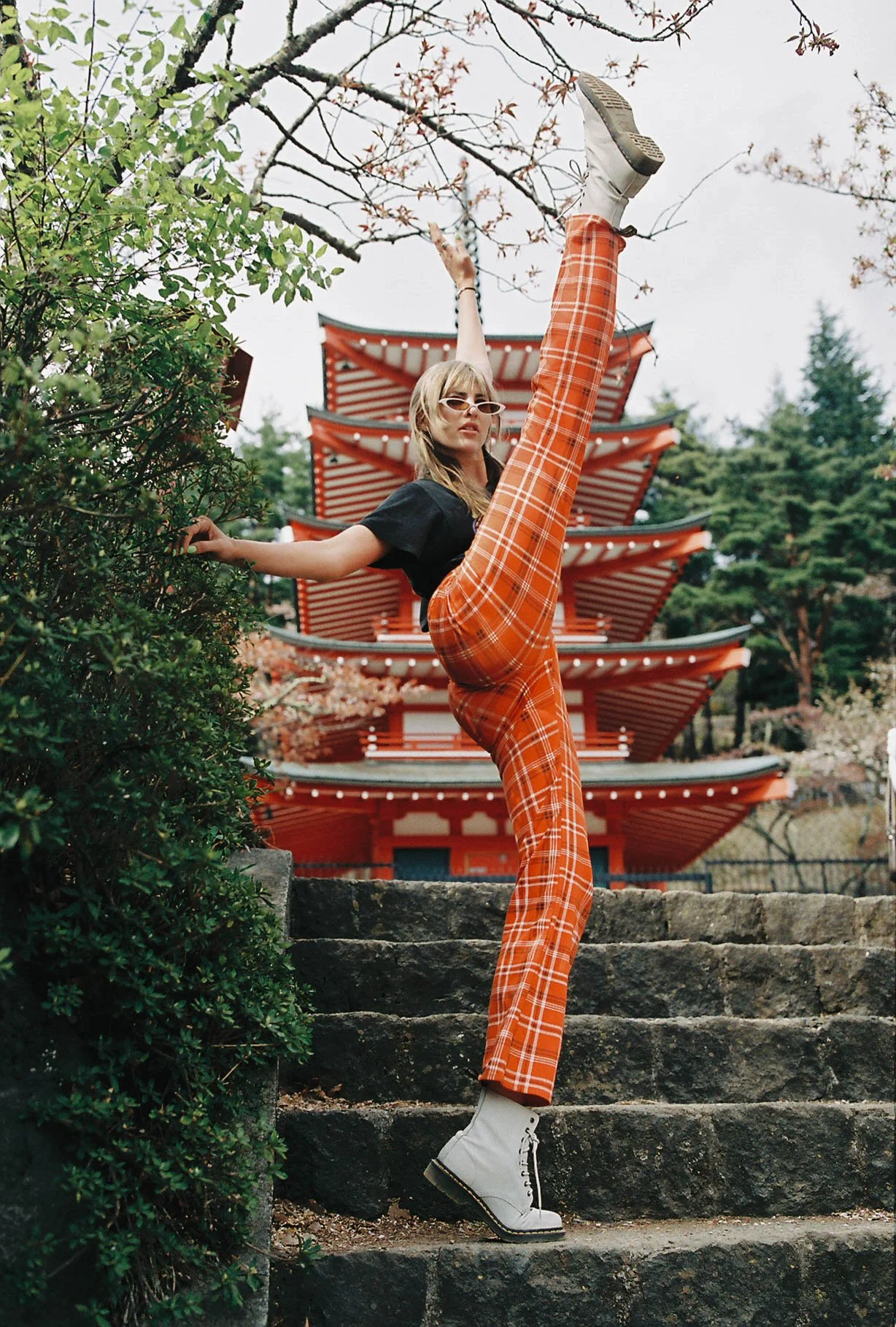 A woman standing on stone steps in front of a red pagoda, striking a high kick, wearing orange plaid pants, a black t-shirt, white boots, and sunglasses, with trees in background.