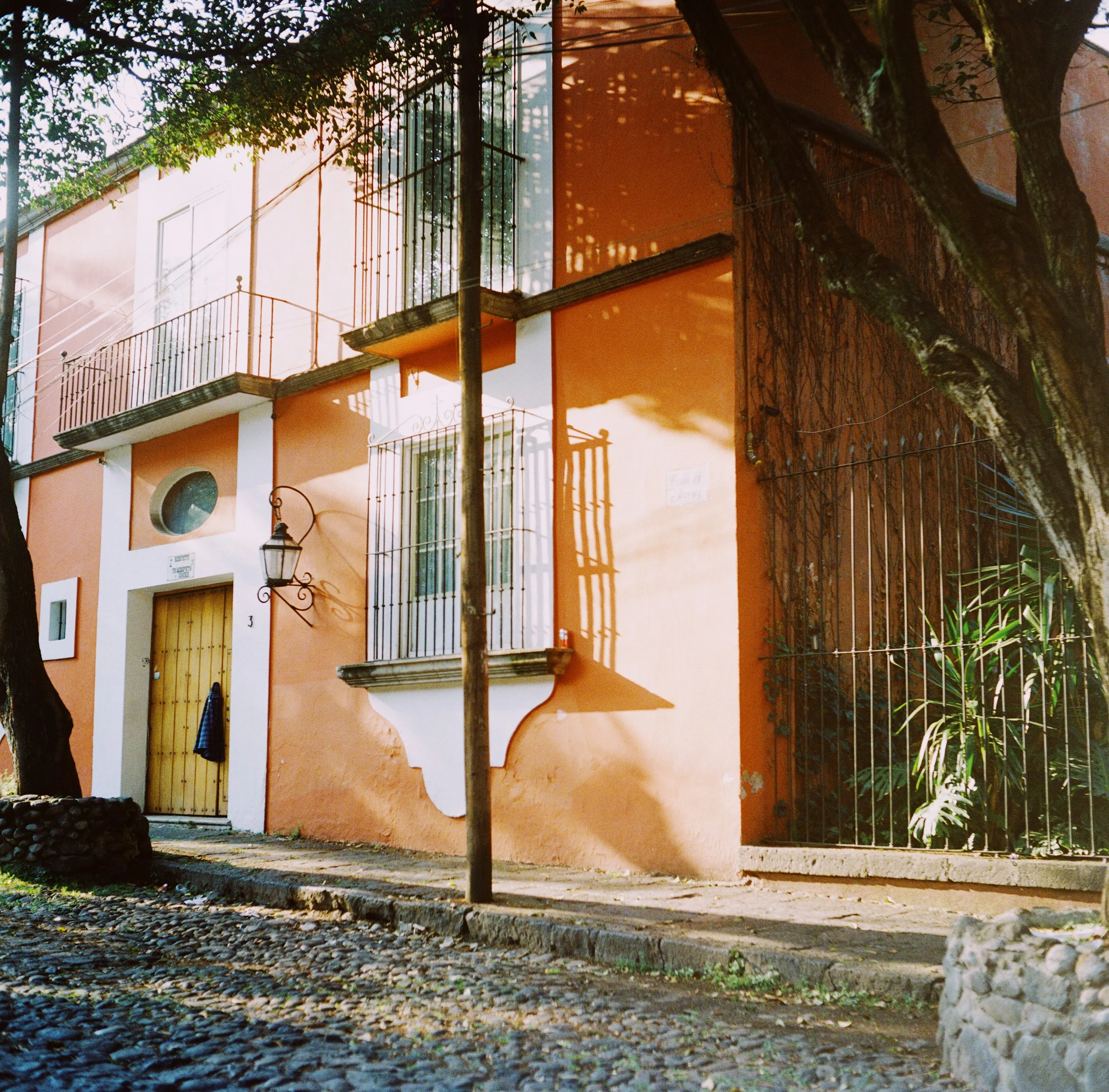 A colorful orange building with a wooden front door, black wrought iron balconies, a streetlamp, and trees casting shadows on the façade.