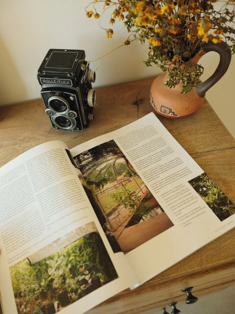A vintage Rolleiflex camera, an open magazine with garden photos, and a flowered vase on a wooden surface.