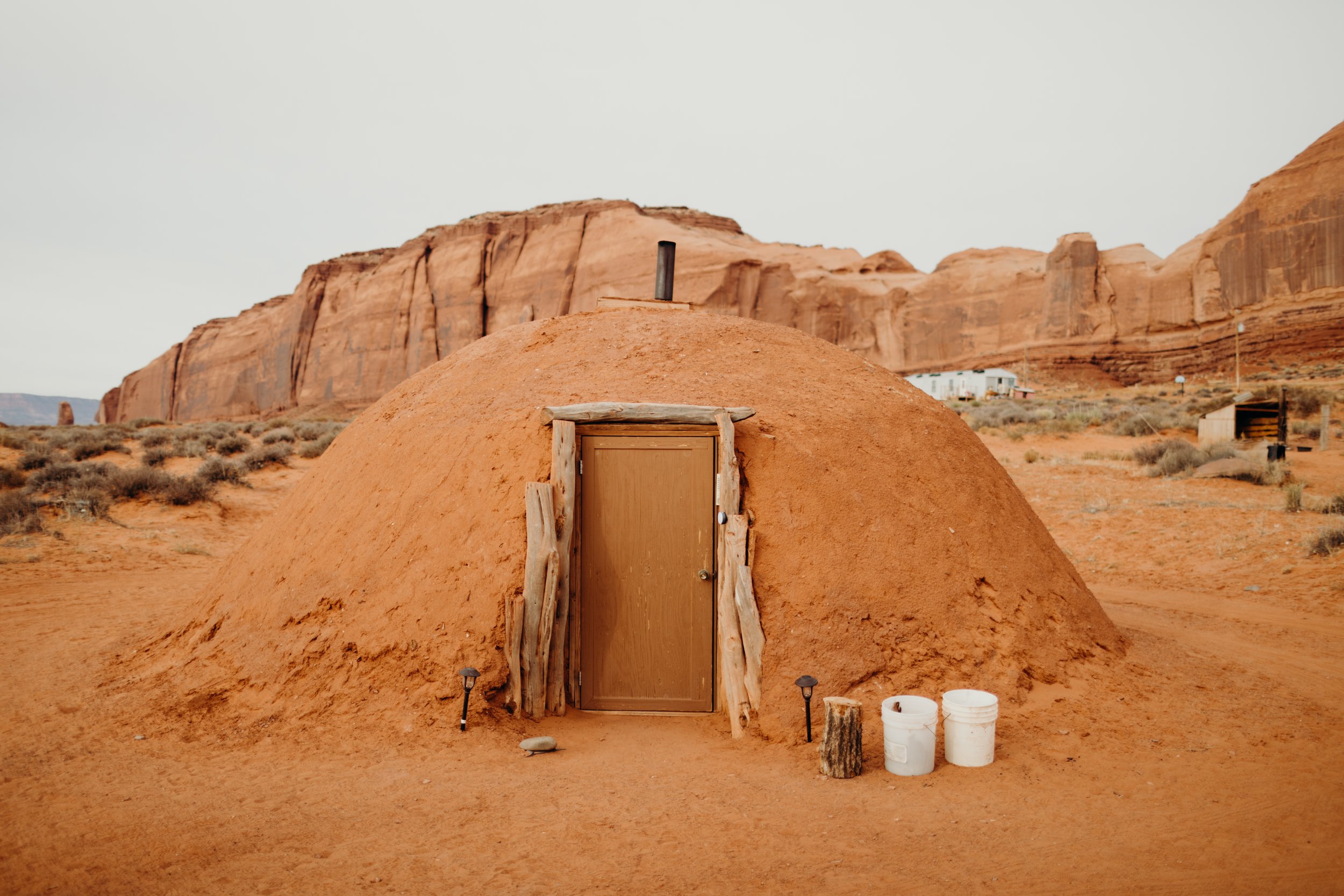 A small adobe-style dwelling in a desert landscape with red rock formations in the background. It has a wooden door framed with driftwood and a chimney on top. There are buckets, a log, and small lamps nearby.