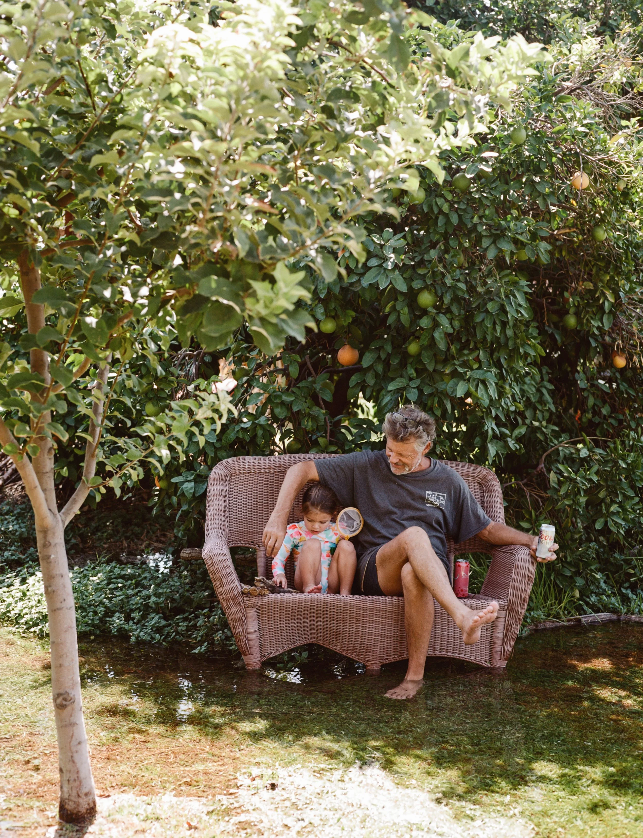 An elderly man and a young girl sitting on a wicker outdoor couch in a backyard with lush green bushes and trees. The man has gray hair and a beard, holding a soda can, and the girl is looking down, playing with something. There is a reflection of su
