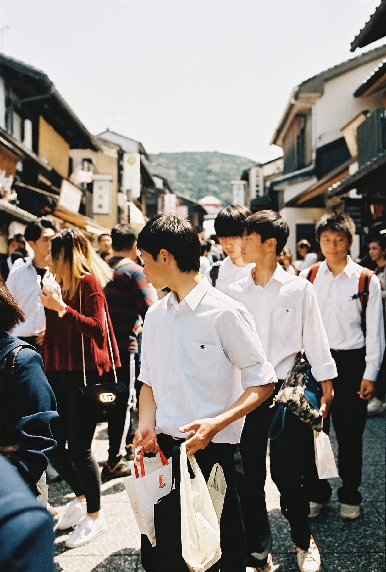Crowd of people walking on a busy street with traditional-style buildings, some are carrying shopping bags, with a mountain in the background.