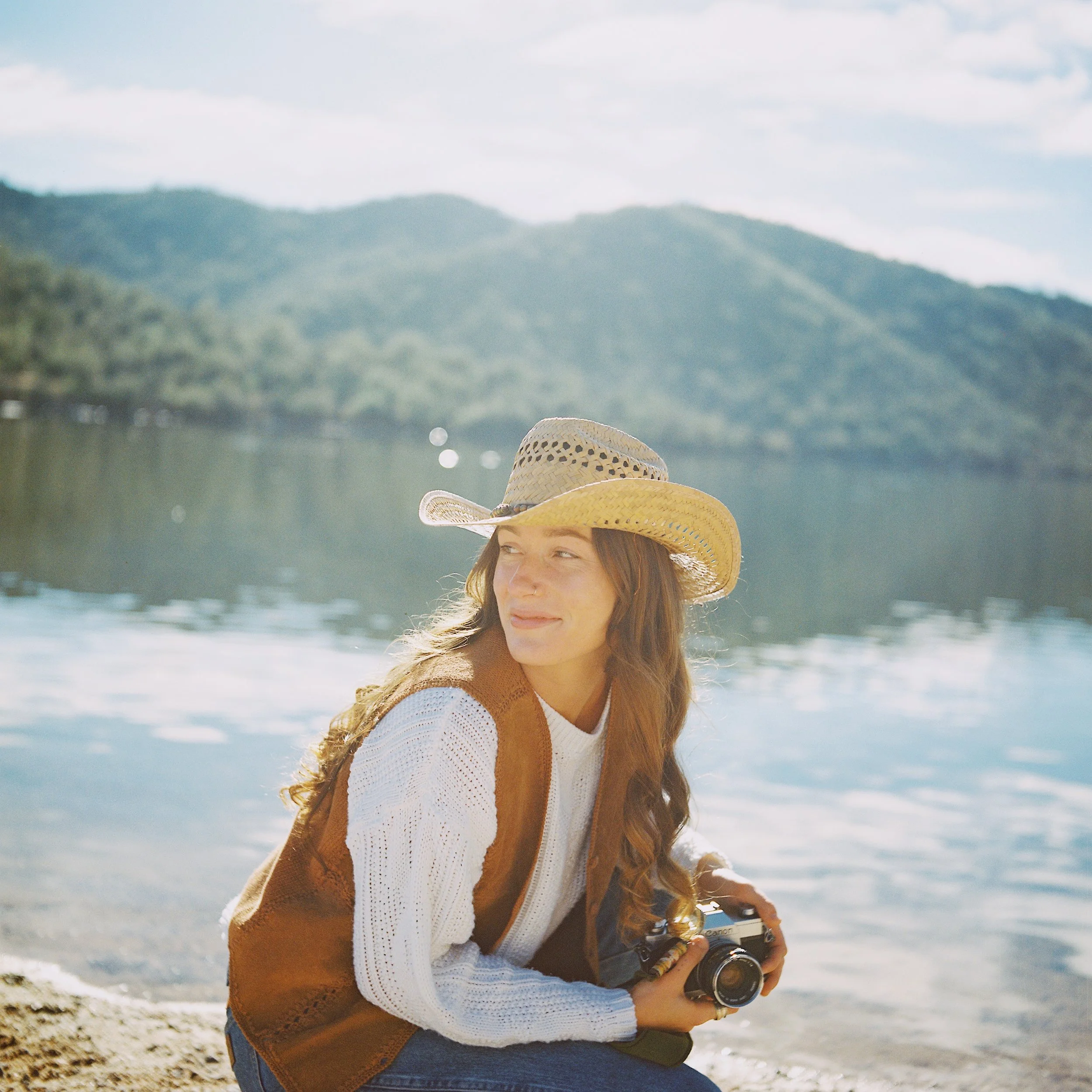 A woman with long hair wearing a straw hat, white knit sweater, and brown vest, holding a camera while sitting by a lake with mountains in the background.