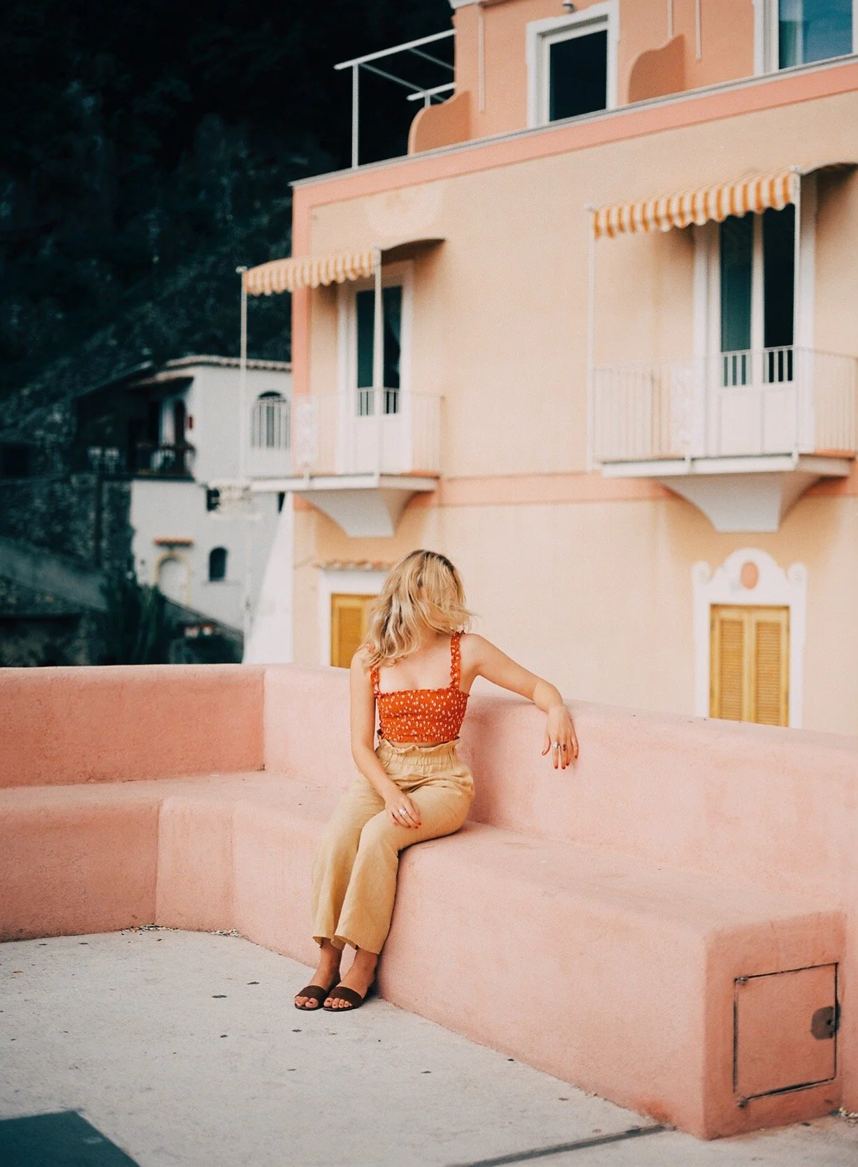 A woman with blonde hair, wearing a red patterned tank top and beige pants, sitting on a pink concrete ledge in front of a pastel-colored building with balconies and awnings.