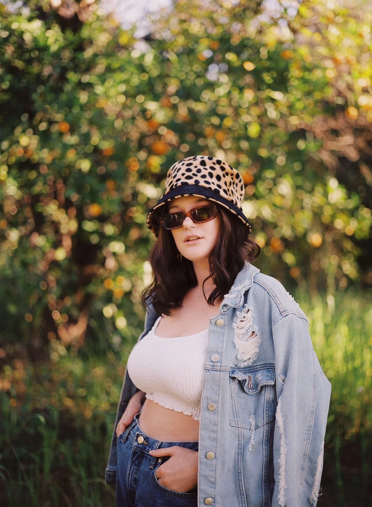 A woman with dark wavy hair wears a leopard-print bucket hat, sunglasses, a distressed denim jacket, a white crop top, and jeans, standing outdoors in a green, sunlit setting.