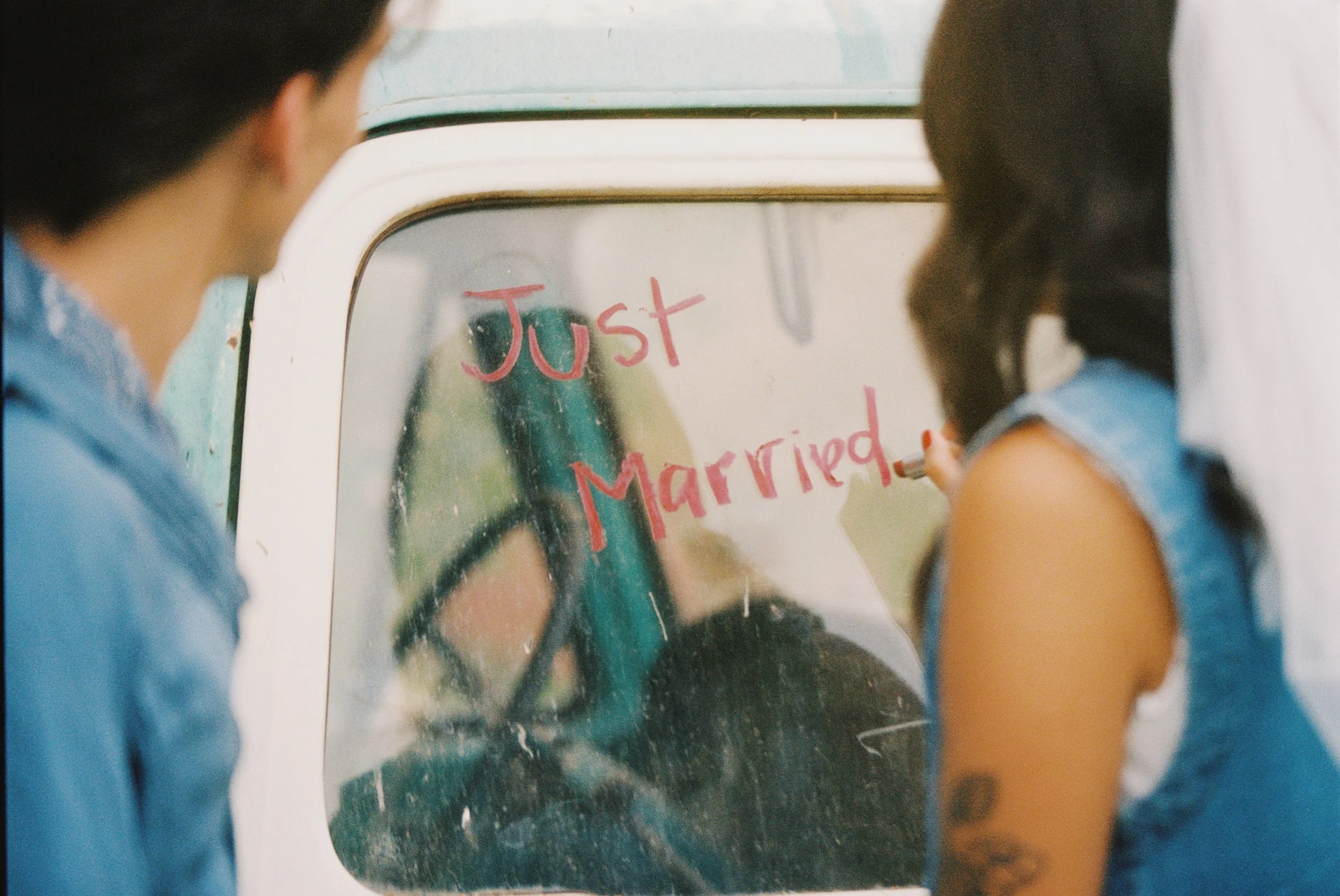 Two people writing 'Just Married' on a glass window with a marker, with an old vehicle in the background.