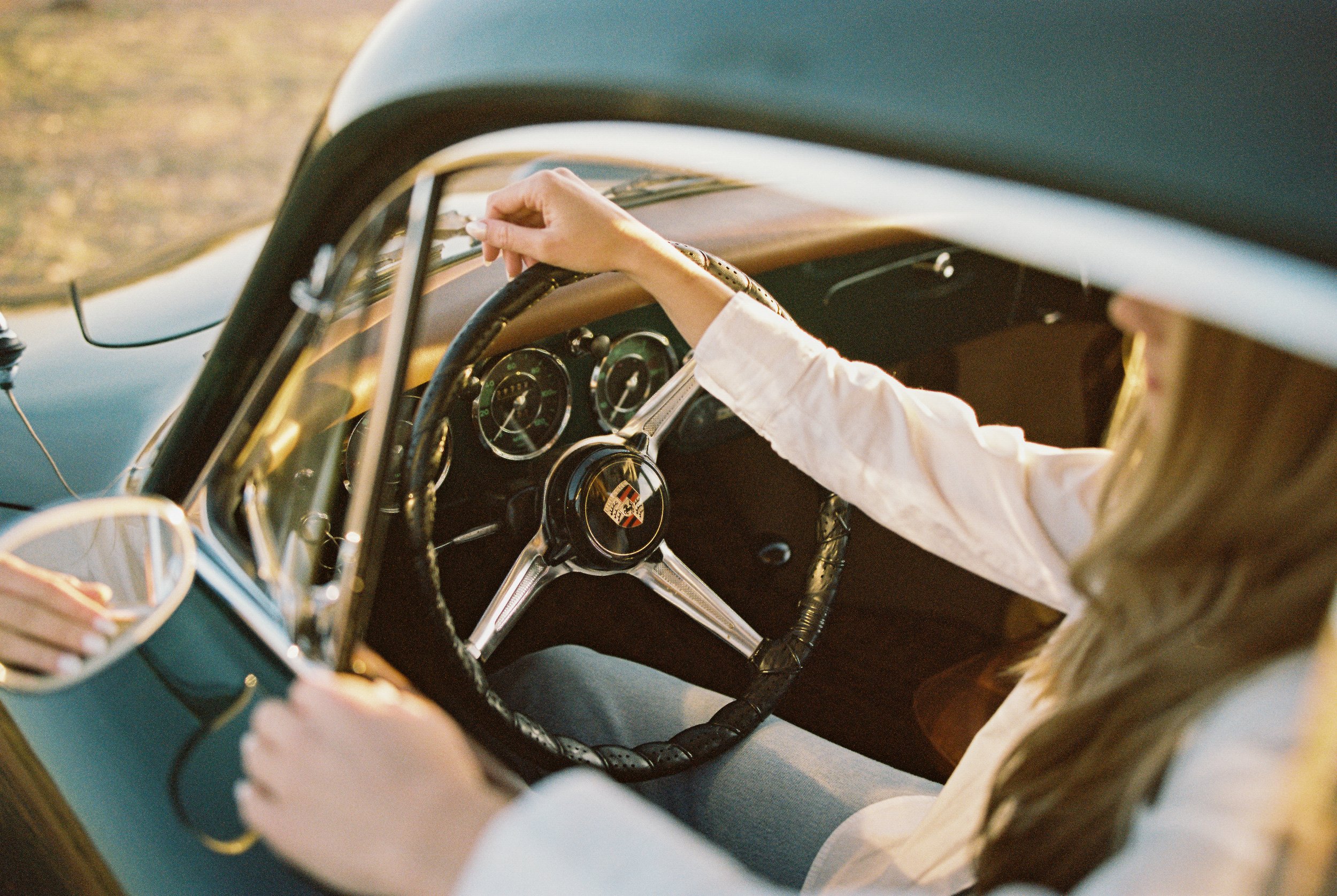 Close-up of a person with long blonde hair driving a vintage car, holding the steering wheel. The dashboard includes classic gauges, and the person is wearing a white long-sleeve shirt. The image is taken from the passenger side with sunlight illumin