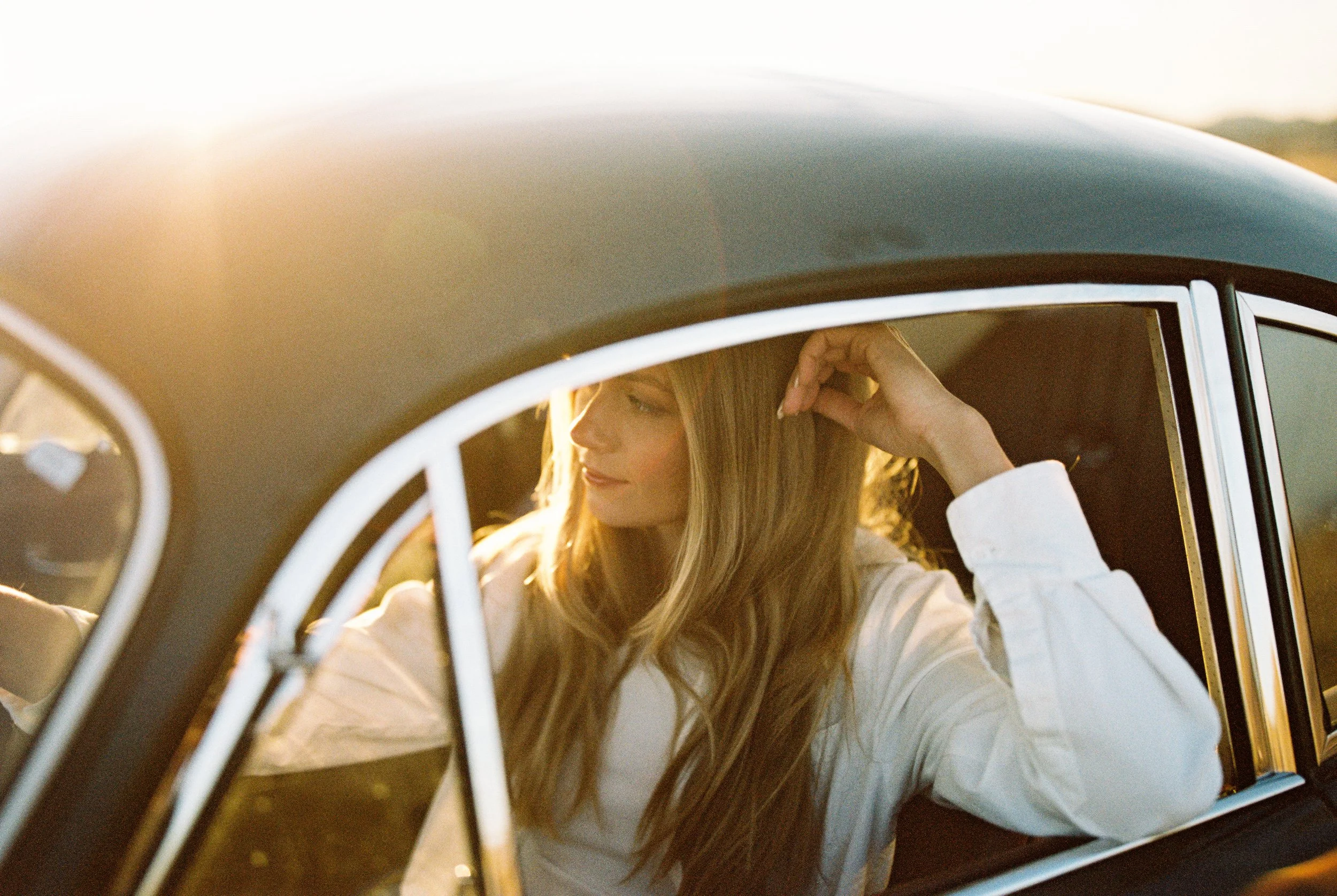 A woman with long blonde hair sitting inside a vintage car, with sunlight streaming in through the window.