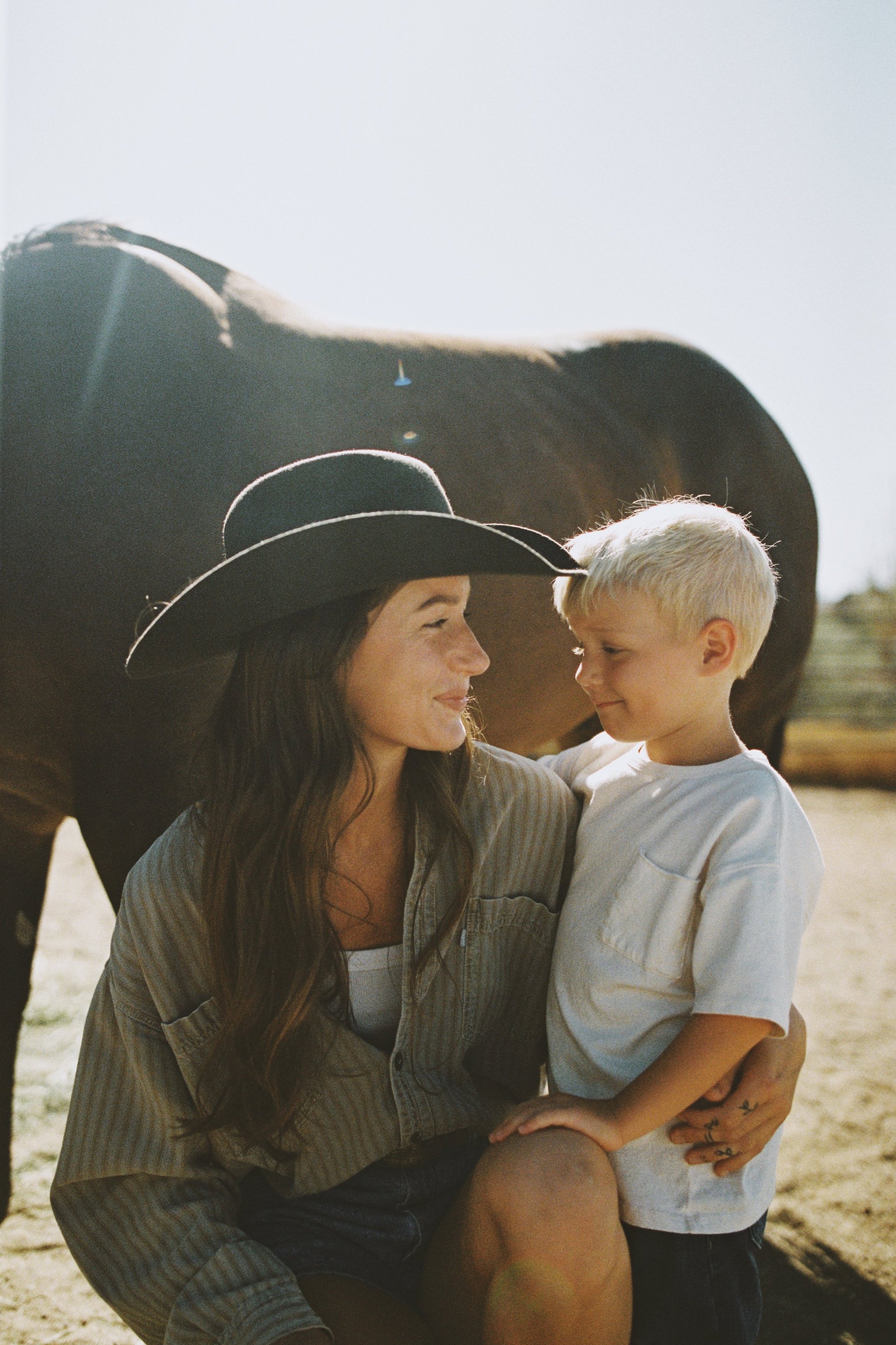 A woman with a large black hat and a young boy in a white shirt sharing a tender moment outdoors at a farm with a horse in the background.