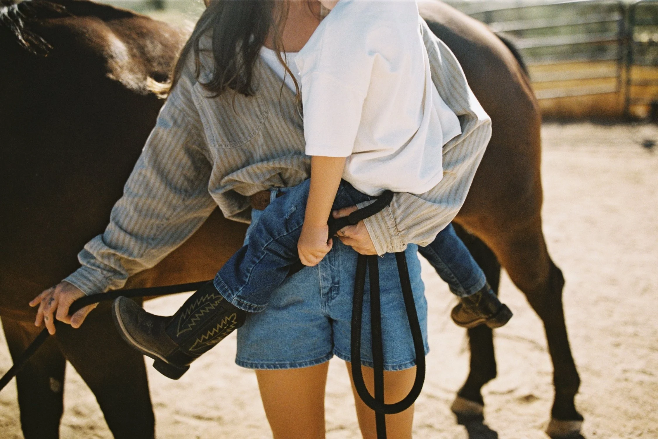 A person in a striped shirt is holding a child, who is wearing a white t-shirt and jeans, while the child is riding on the person's hip near a brown horse in an outdoor setting.