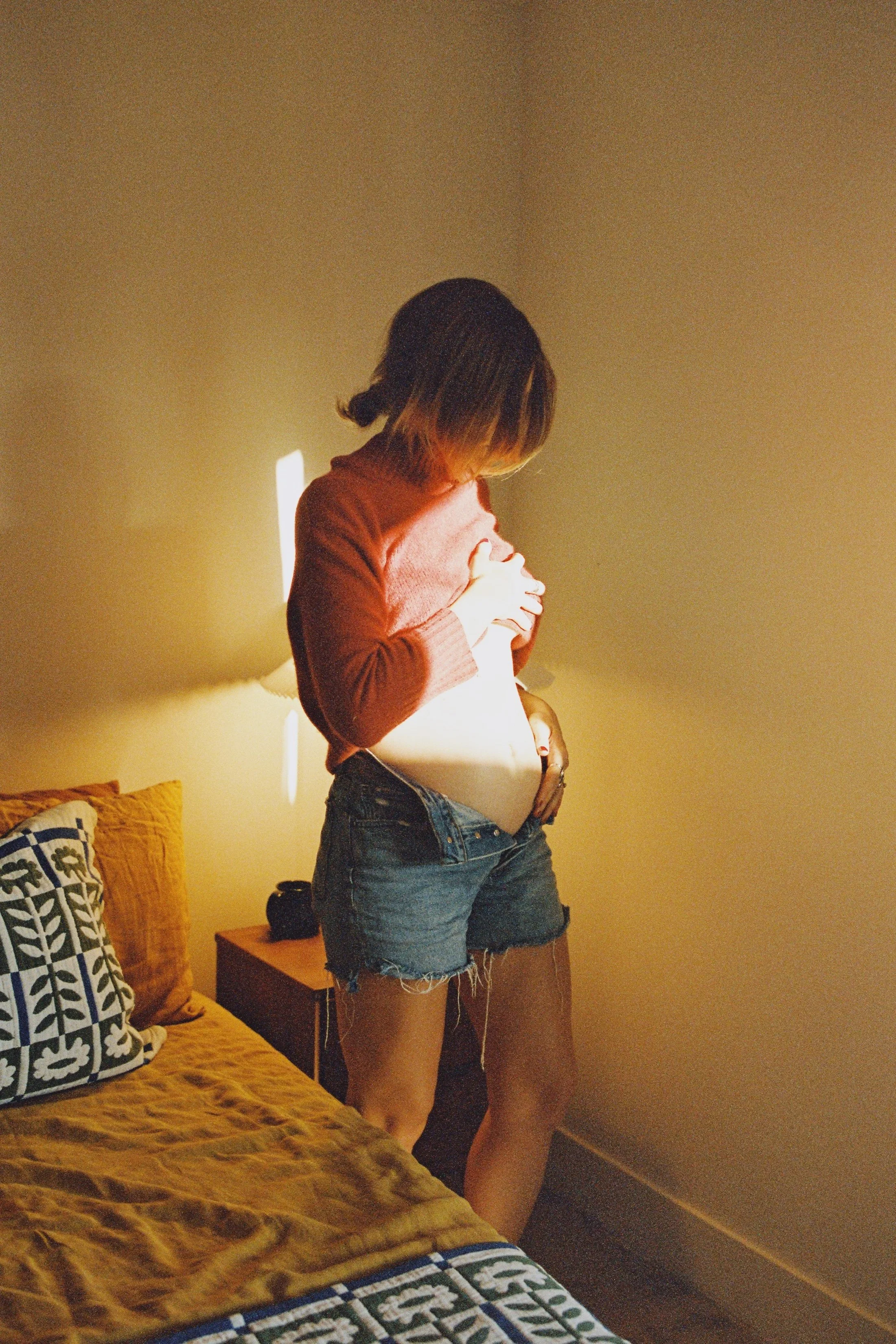 A woman with shoulder-length dark hair, wearing a red long-sleeve shirt and denim shorts, stands in a bedroom. She is lifting her shirt to reveal her stomach, holding it with one hand, while her other hand is on her lower abdomen. The room has warm l