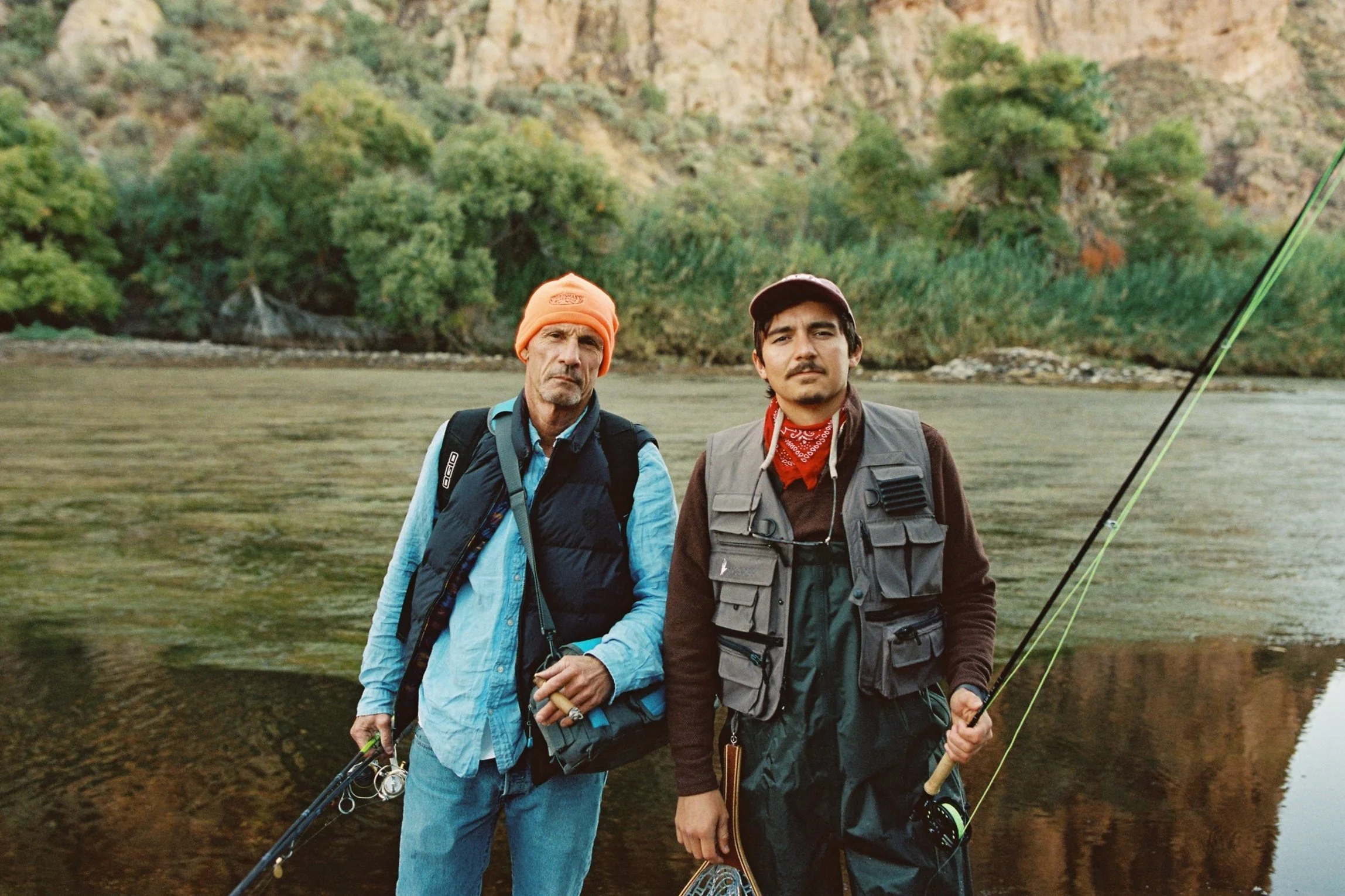 Two men standing by a river, holding fishing rods, with a wooded hillside in the background.
