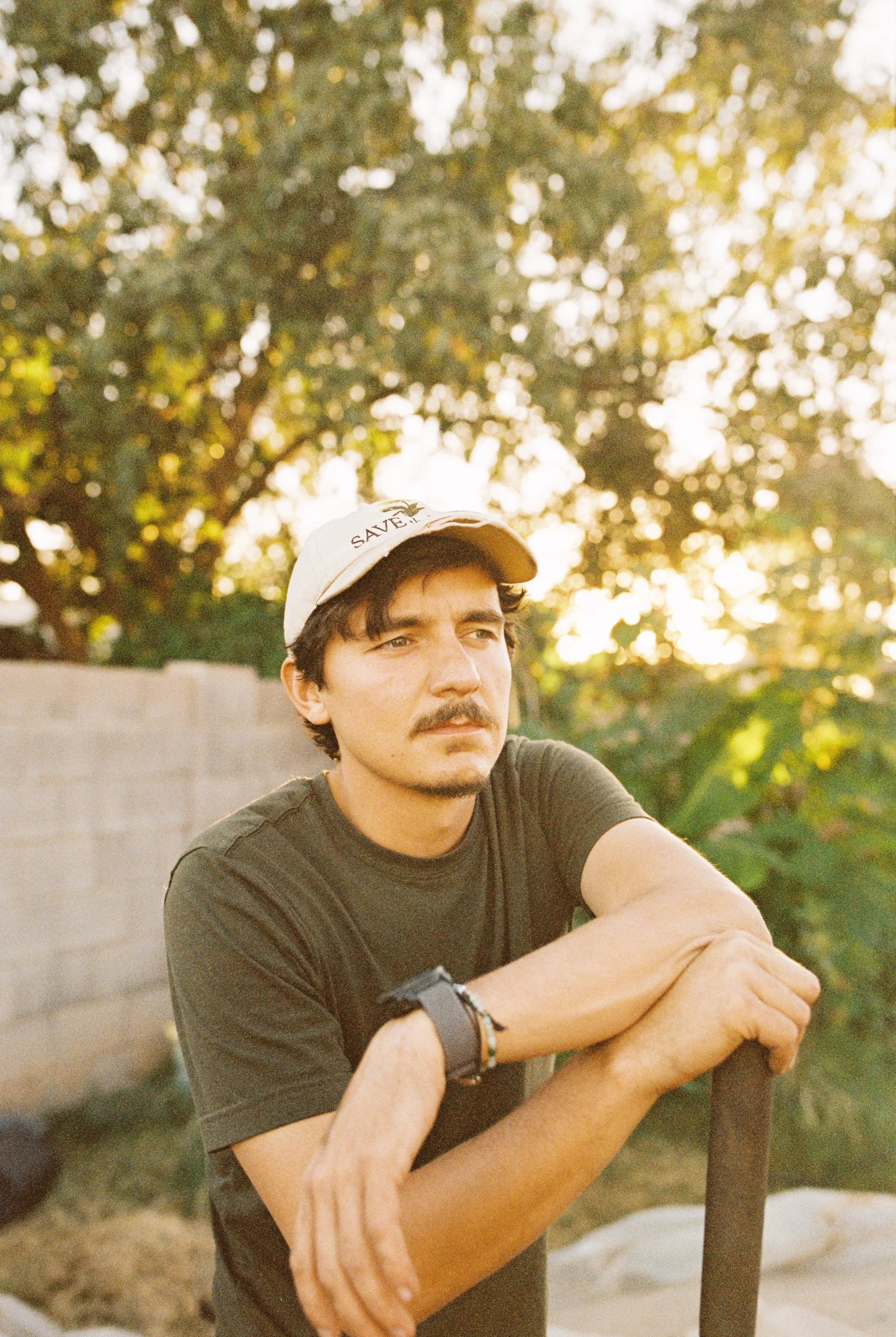 A man with a mustache and dark hair, wearing a beige cap and a dark T-shirt, sits outdoors during sunset, resting his arms on a shovel.