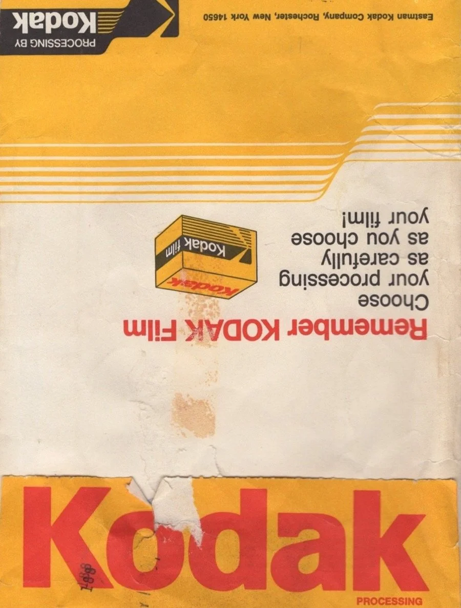 A crumpled and worn Kodak film packaging box with the words 'Remembear Kodak film' printed in red and yellow on it, with a Kodak logo on a small box illustration. The box has a yellow, white, and black color scheme.