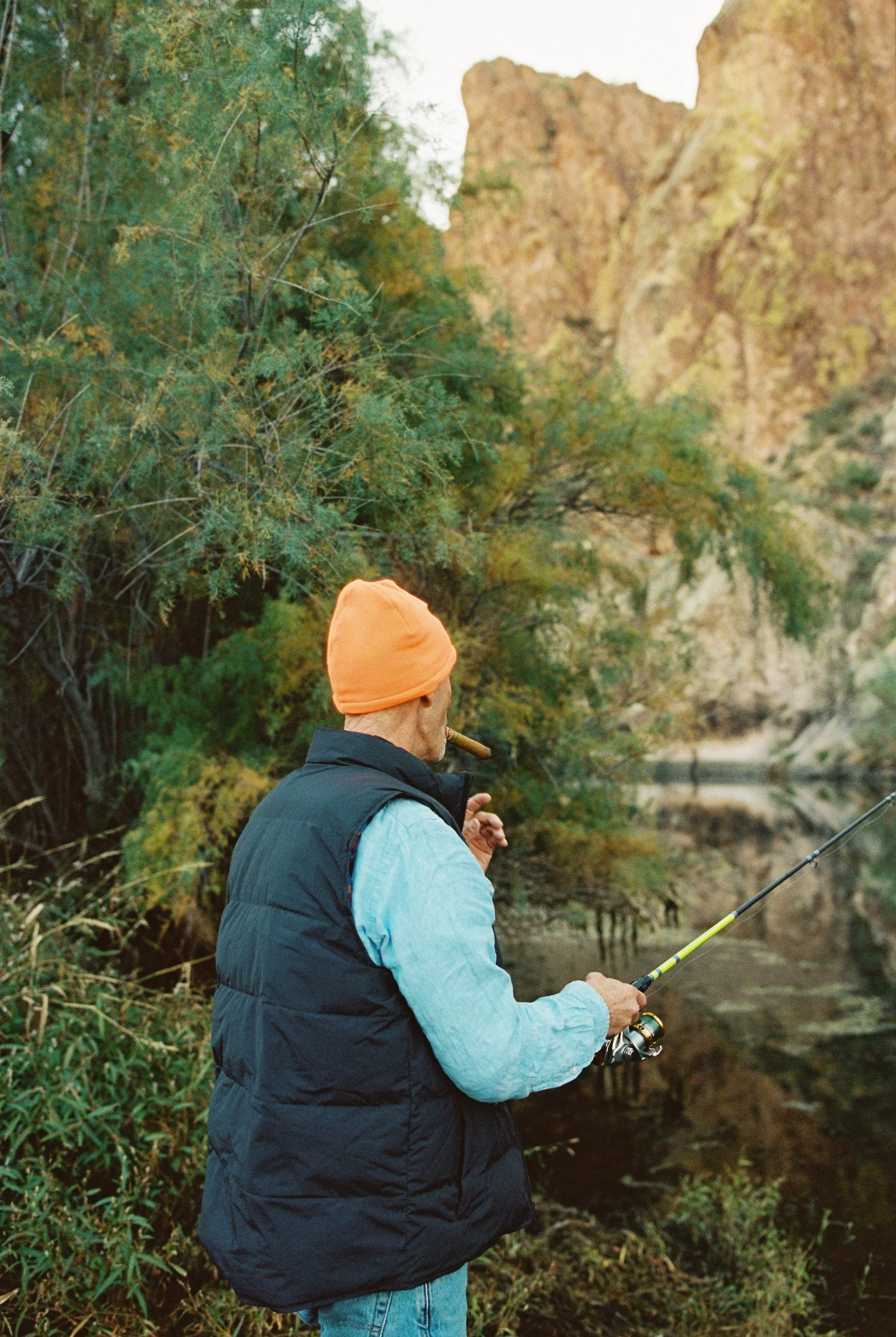 A man fishing by a river with rocky cliffs and trees in the background, wearing an orange beanie, blue denim shirt, and black vest, smoking a cigar.