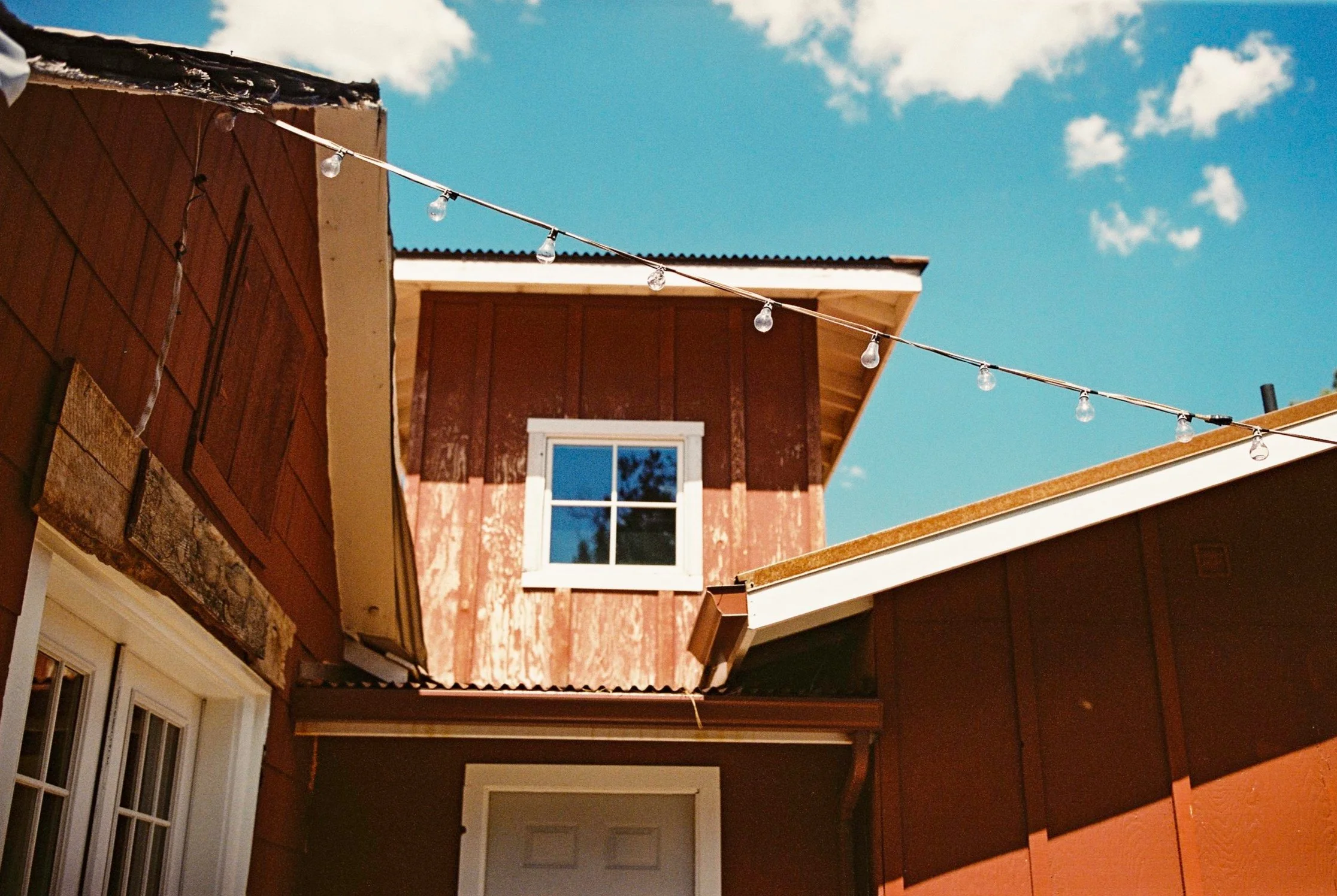 Close-up of a rustic house with a weathered red wooden exterior, a white-framed window, and a string of outdoor lights against a clear blue sky.