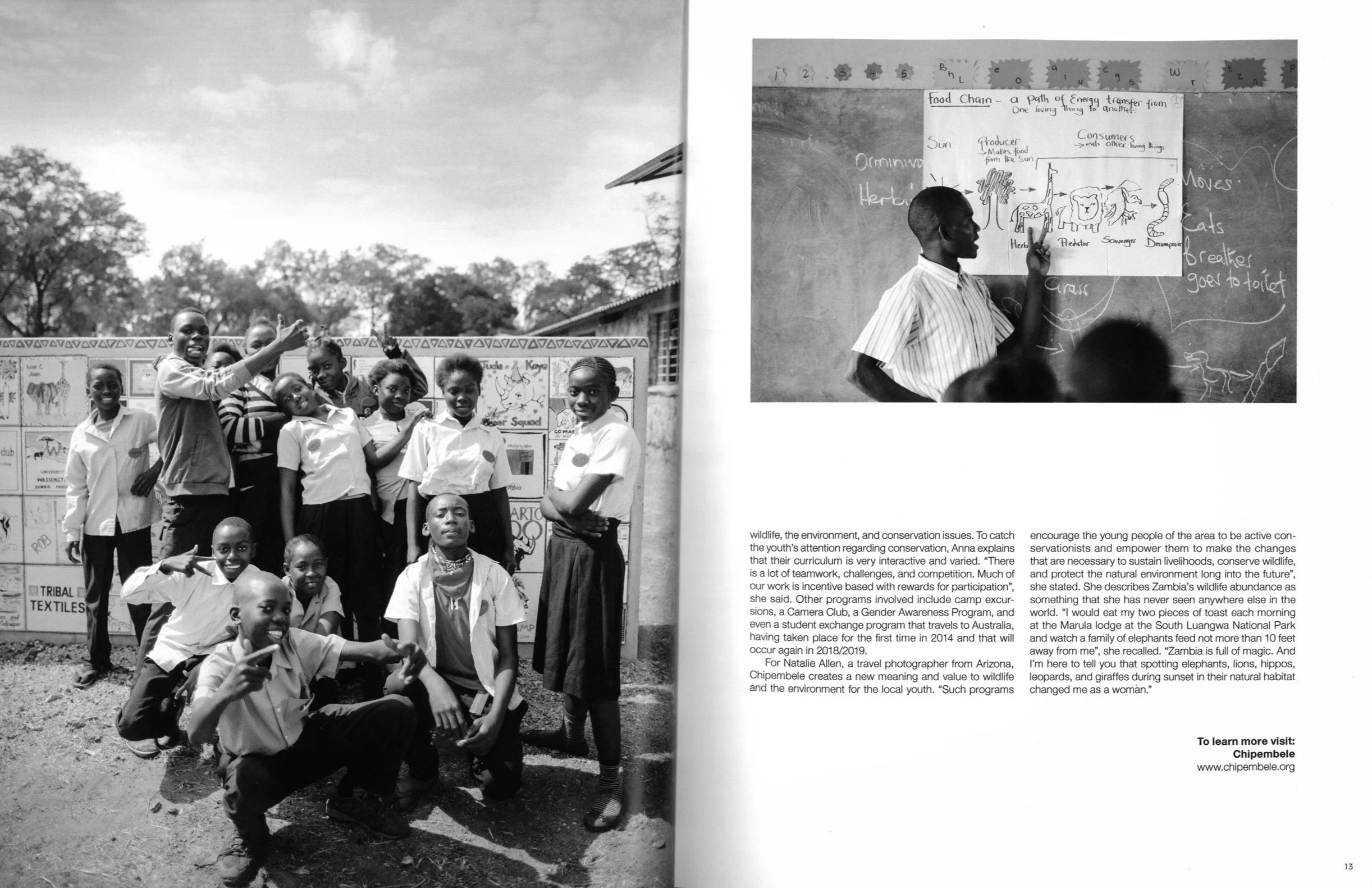 Group of children and a teacher outdoors in front of a display board, school building in background, some children making peace signs, teacher and students posing for the camera.