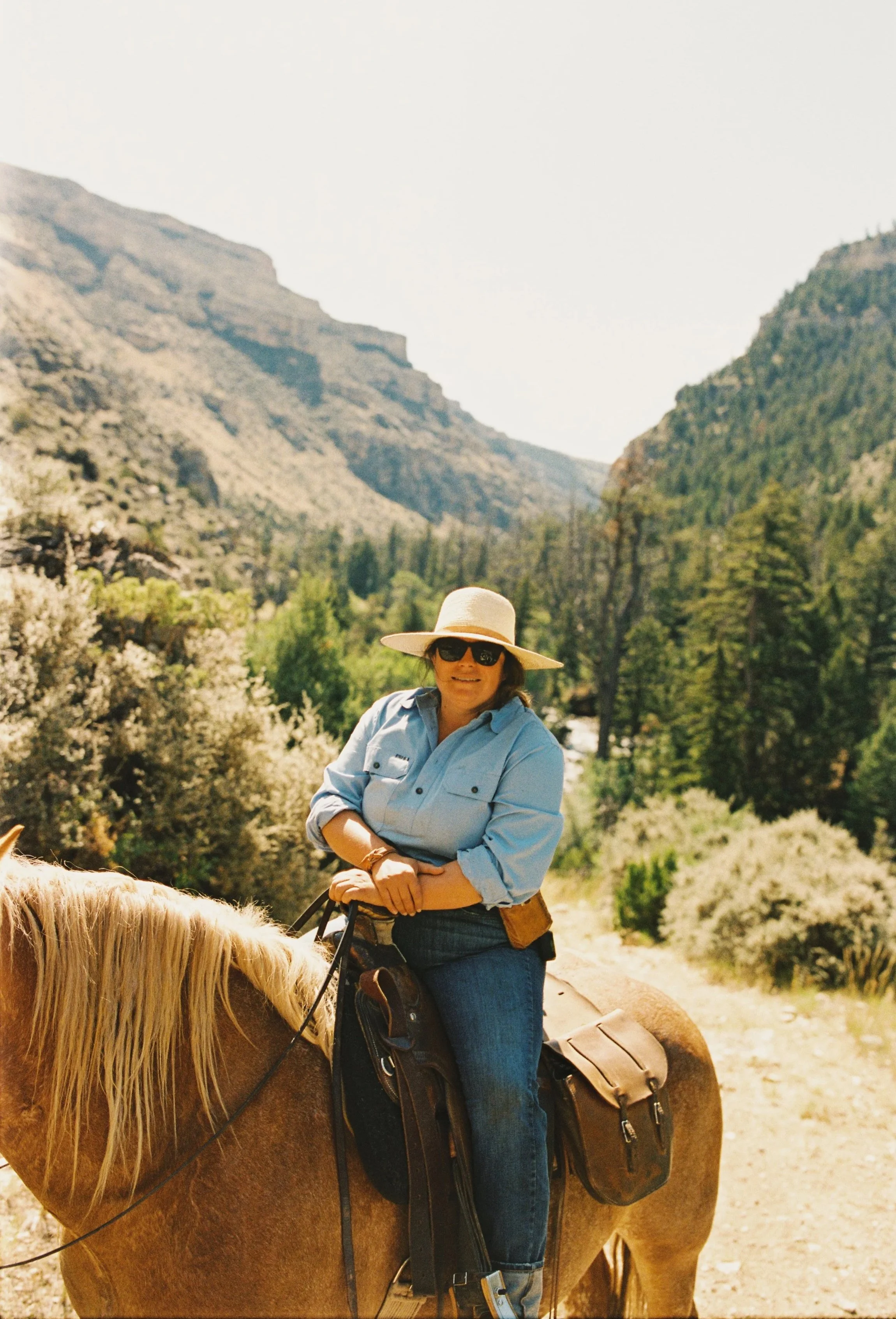 A woman wearing a wide-brimmed hat, sunglasses, a light blue shirt, and jeans is sitting on a horse in a mountainous landscape with trees and rocky cliffs.
